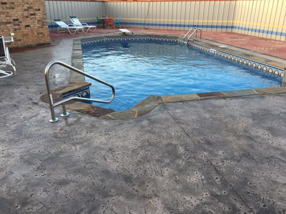 Outdoor swimming pool with a metal handrail, concrete deck, and a brick wall backdrop