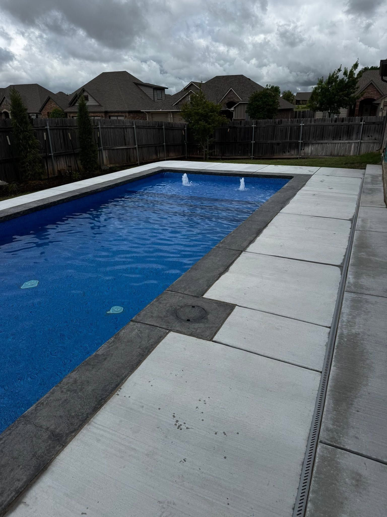 Rectangular backyard swimming pool with blue water and white deck pavers under a cloudy sky