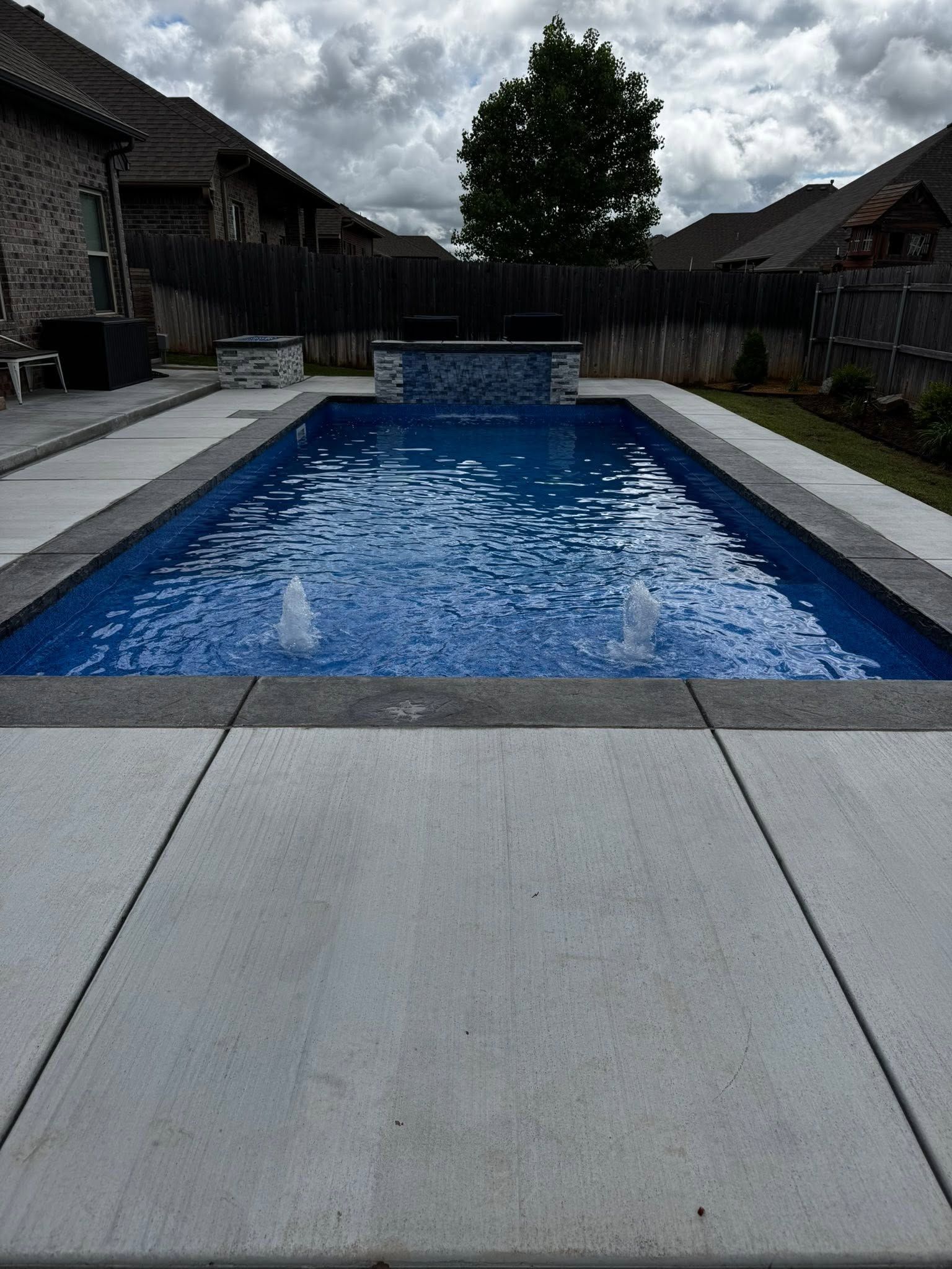 Backyard swimming pool with blue water, gray patio, and cloudy sky, framed by houses and a wooden fence