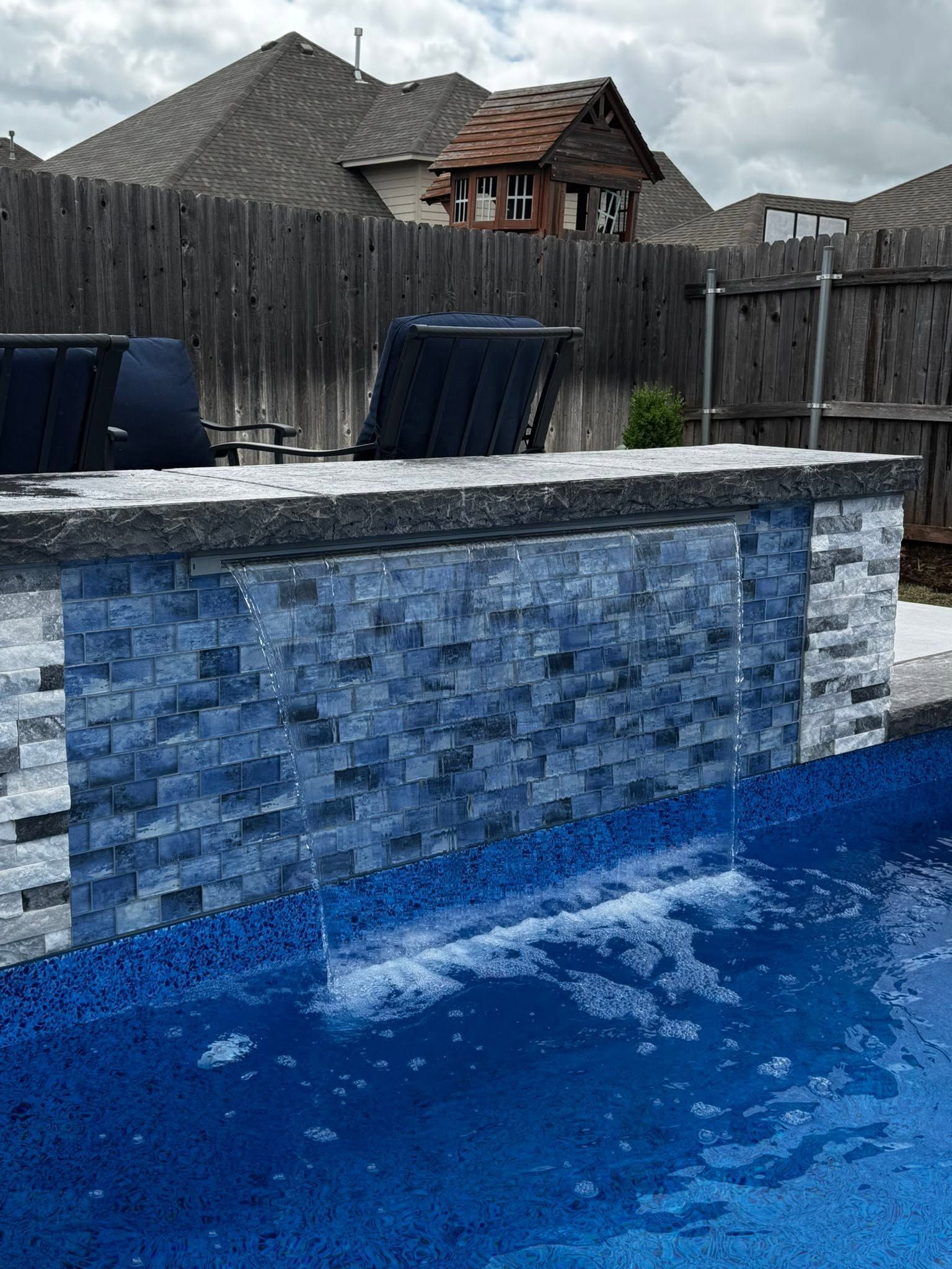 Blue backyard pool with stone ledge, lounge chairs, and a wooden fence in the background