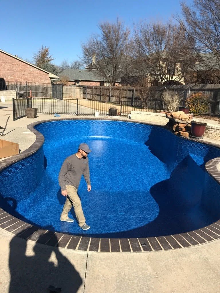 Person walking in an empty blue swimming pool in a fenced backyard during daylight