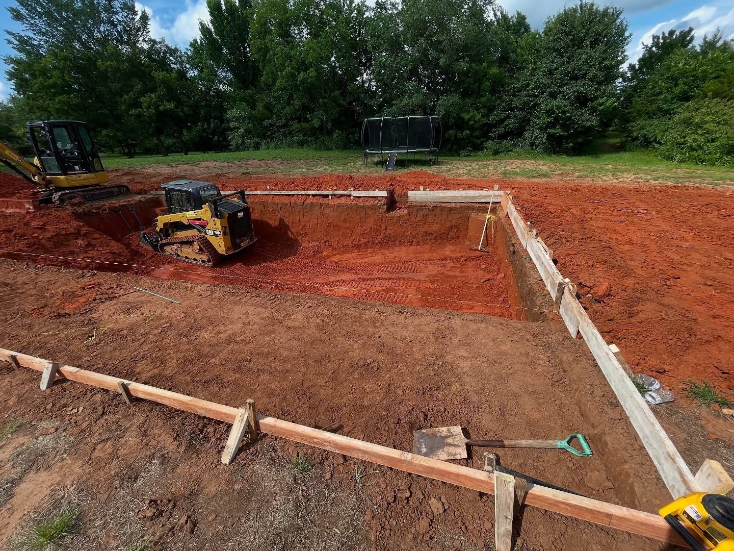 Construction site with excavator and compact loader on red dirt inside a concrete foundation.