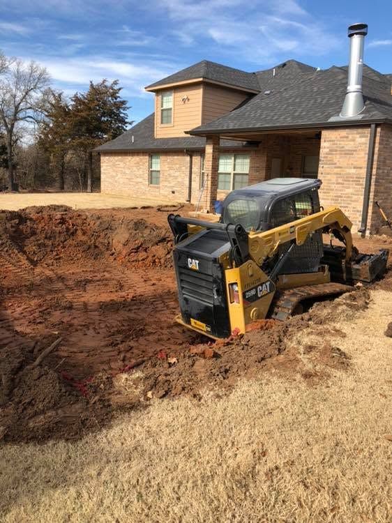 Yellow skid steer excavator digging a large trench beside a brick house and driveway