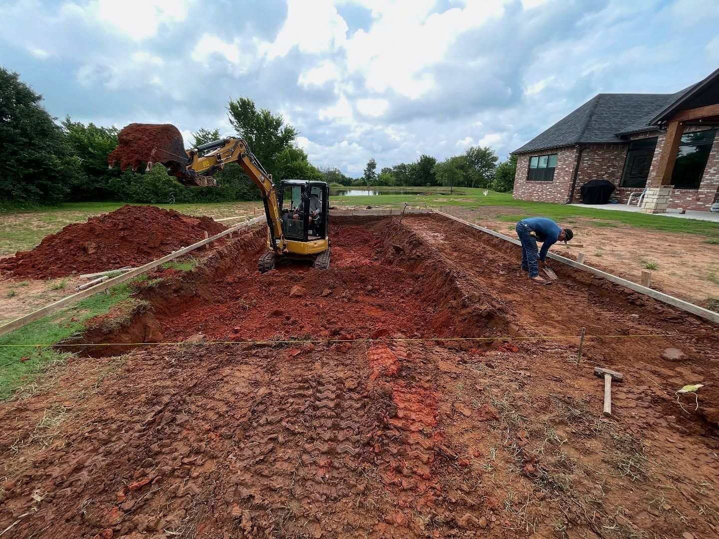 Construction site with excavator and worker grading red dirt near a house under a cloudy sky