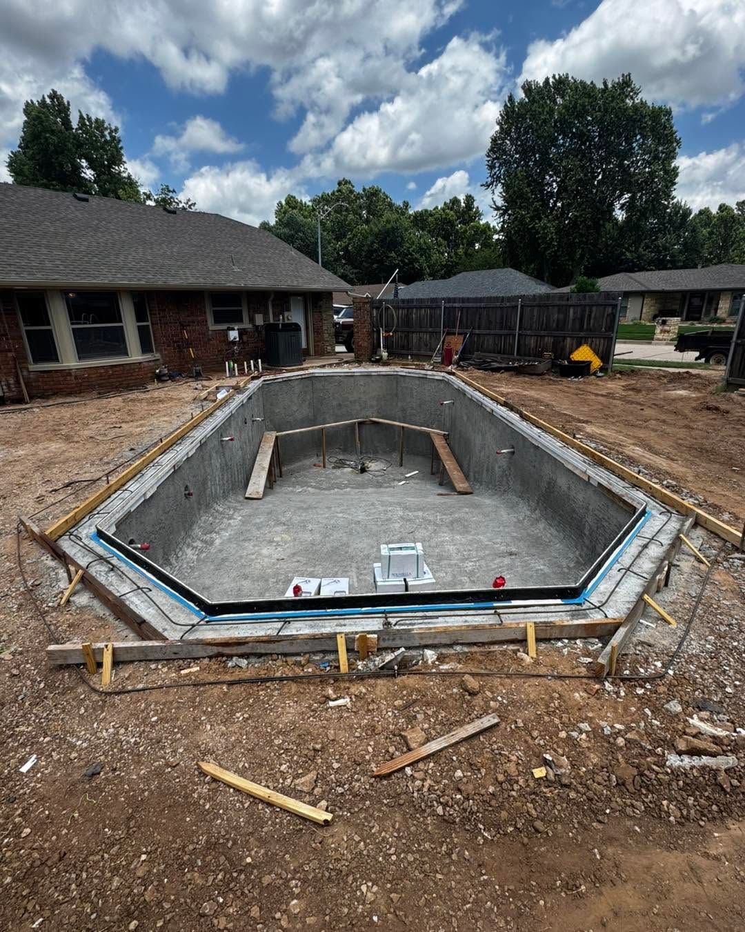 Empty concrete pool under construction in a backyard, surrounded by dirt and construction materials.