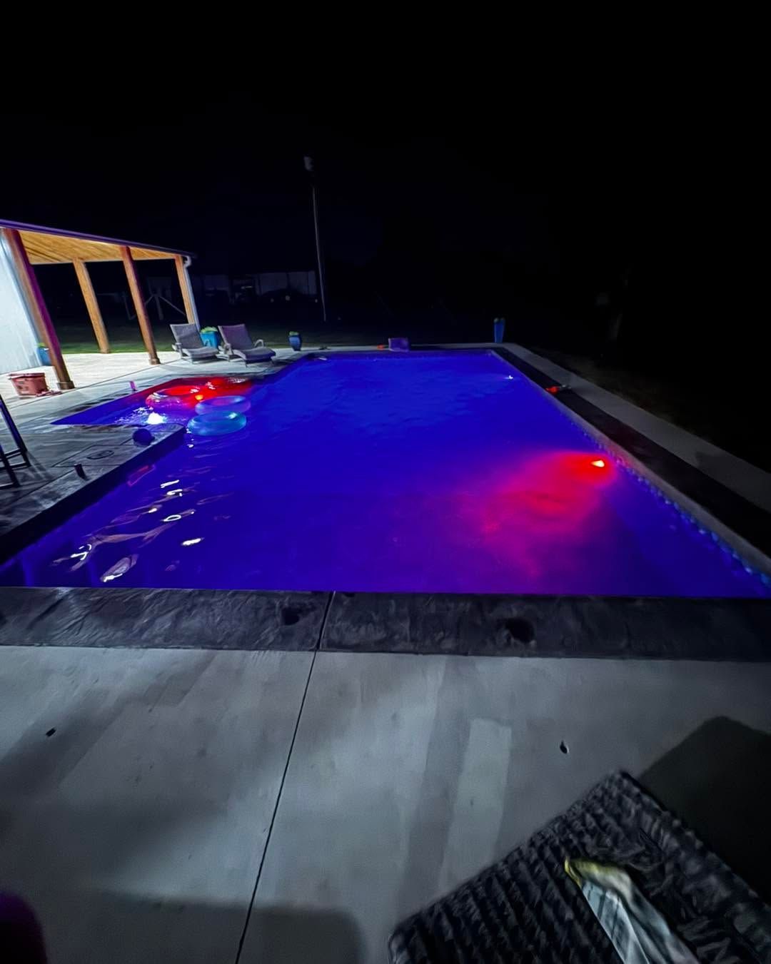 Lit backyard pool glowing blue and purple at night, with a patio and seating area beside it