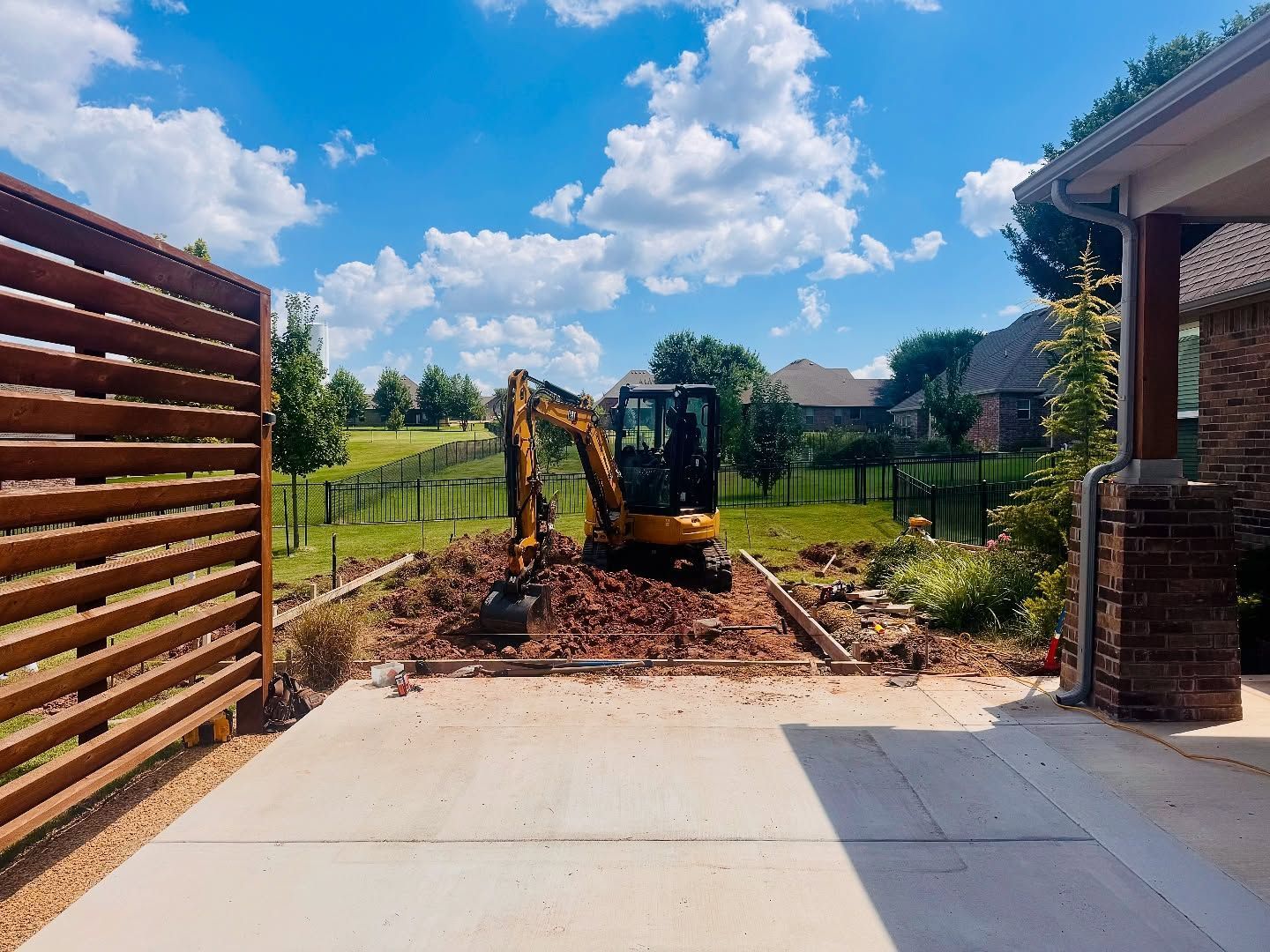 Yellow excavator digging in a driveway opening, with soil piled up under a blue sky.