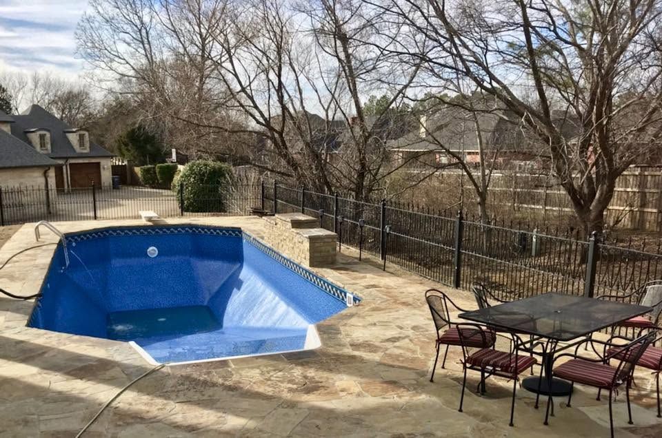 Empty blue in-ground pool beside a patio with outdoor chairs and leafless trees in a backyard