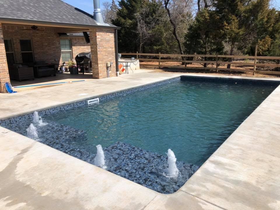 Rectangular backyard swimming pool with stone deck, water jets, and a covered patio beside a brick house