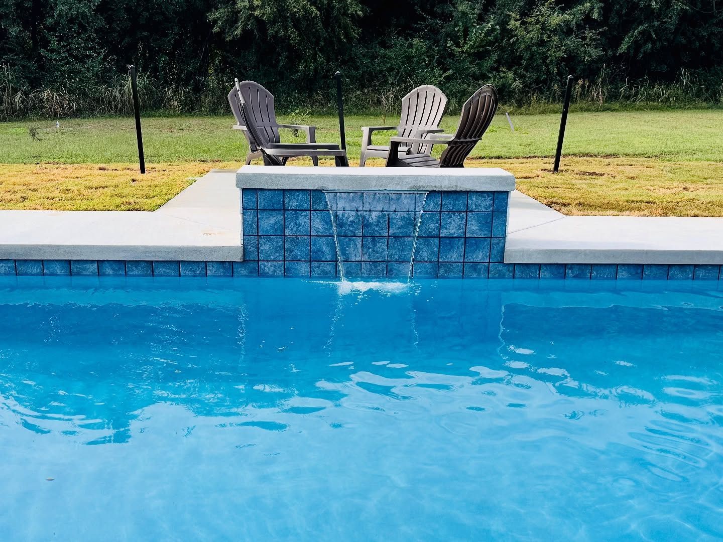 Backyard swimming pool with blue water, stone fountain, and two chairs on the deck