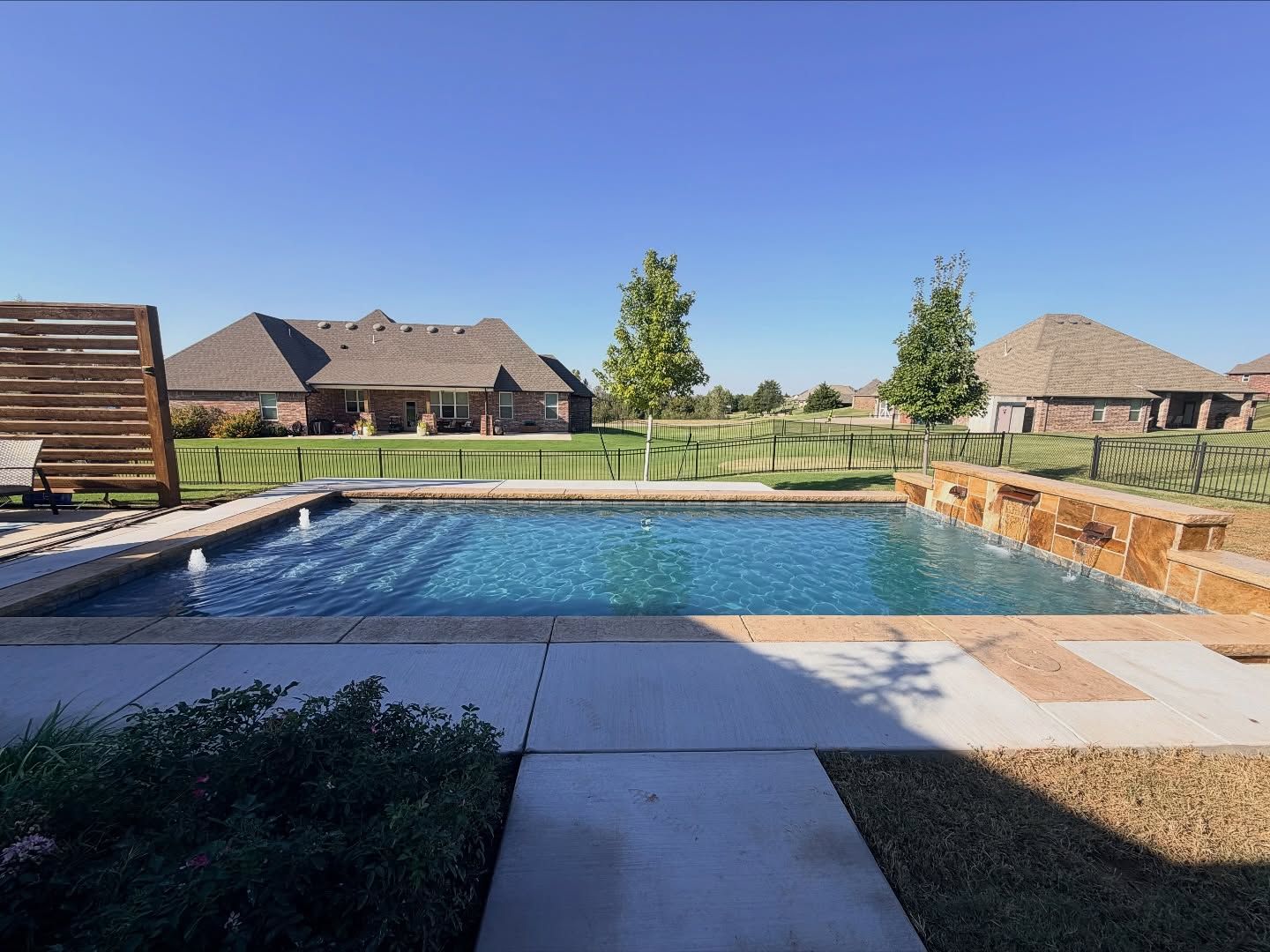Backyard pool with stone deck, patio, and houses under a clear blue sky