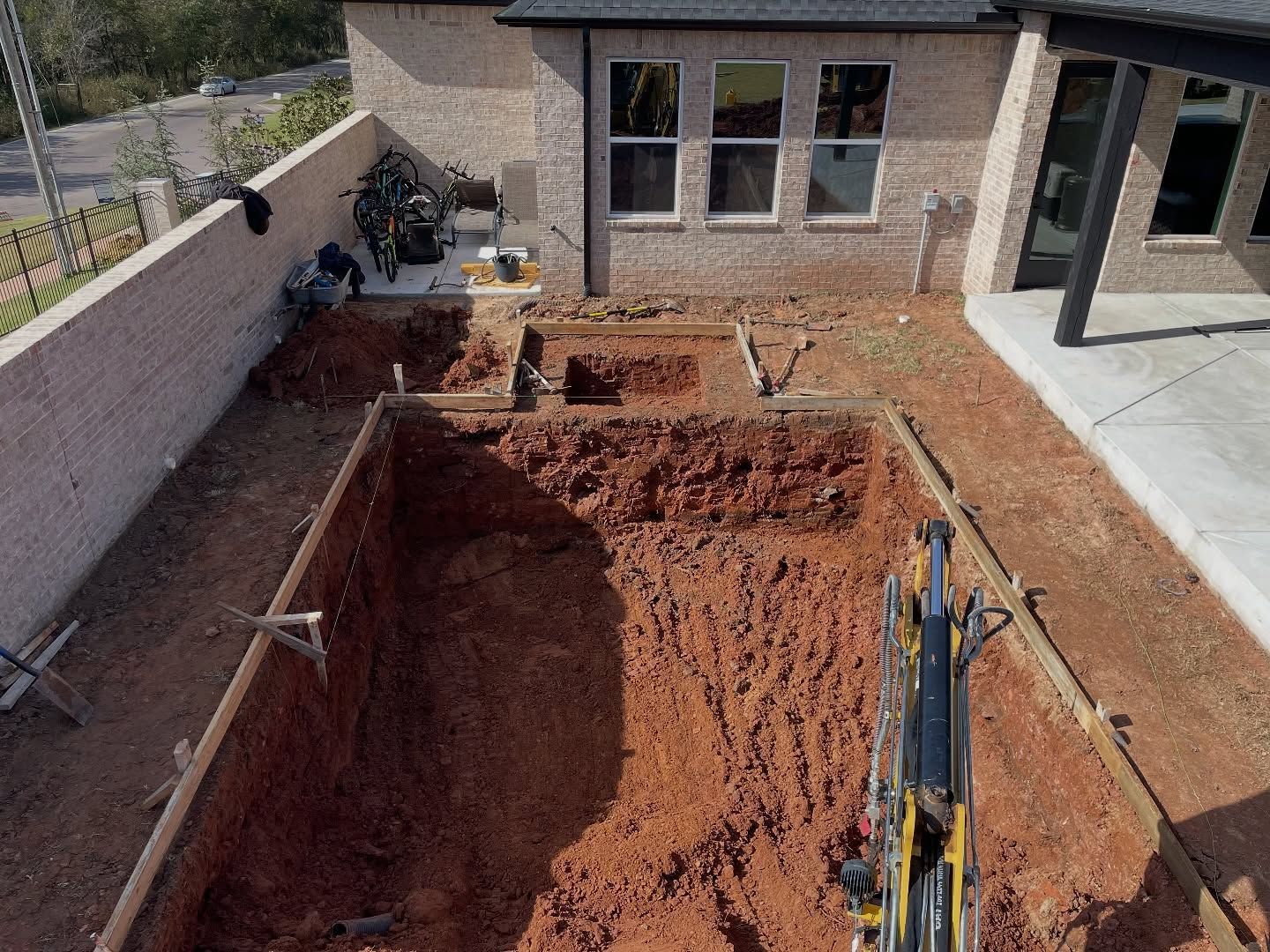 Large red-soil excavation pit beside a brick building, with tools and a ladder on the right edge.