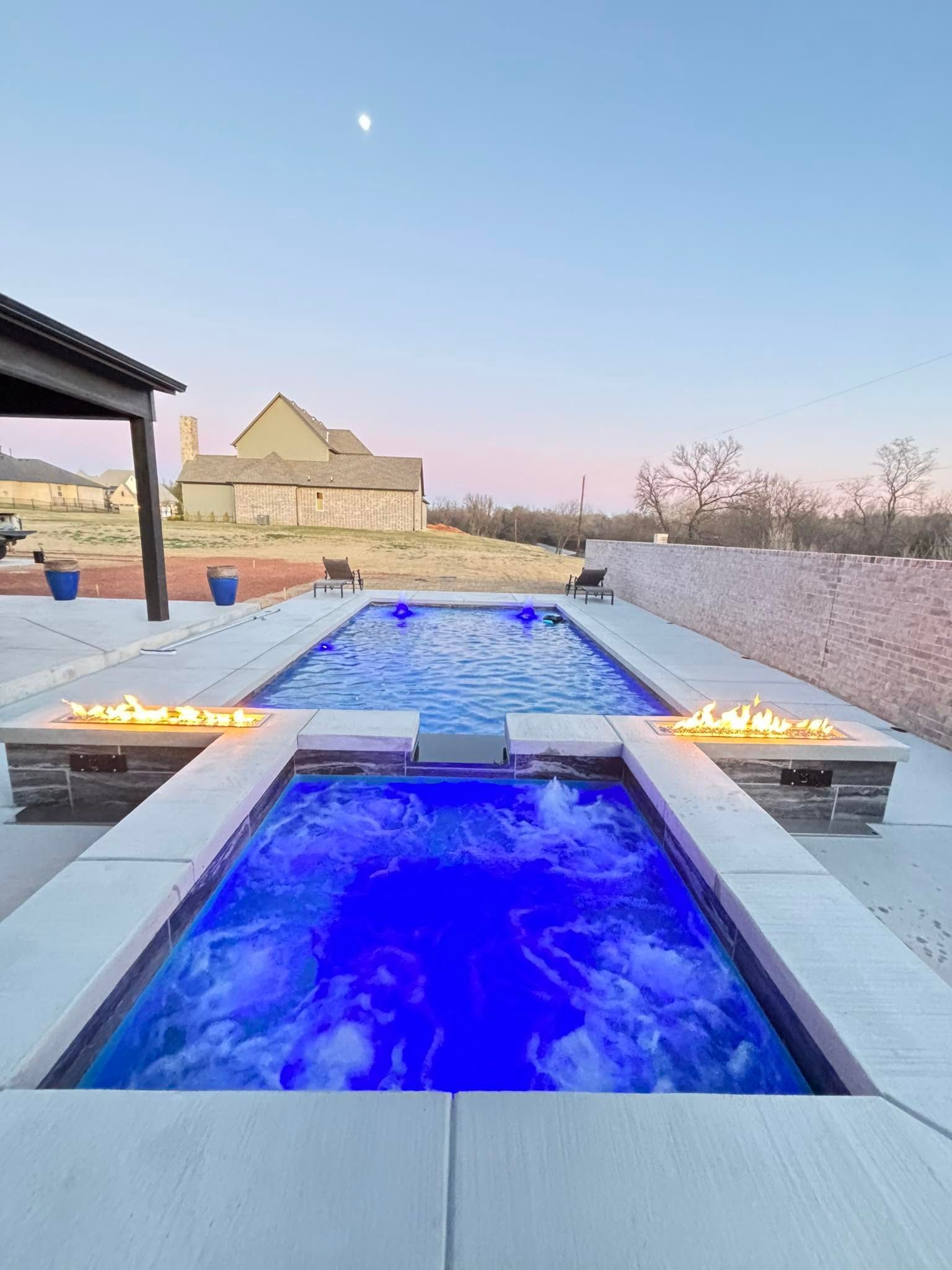 Blue-lit outdoor pool with fire features at dusk, overlooking a stone building and pale sky