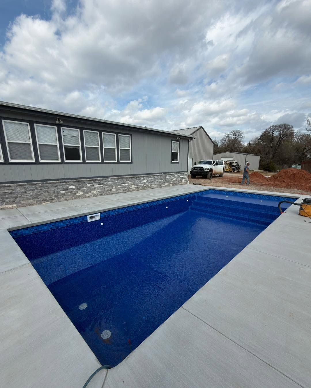 Blue rectangular backyard swimming pool beside a gray house under a cloudy sky