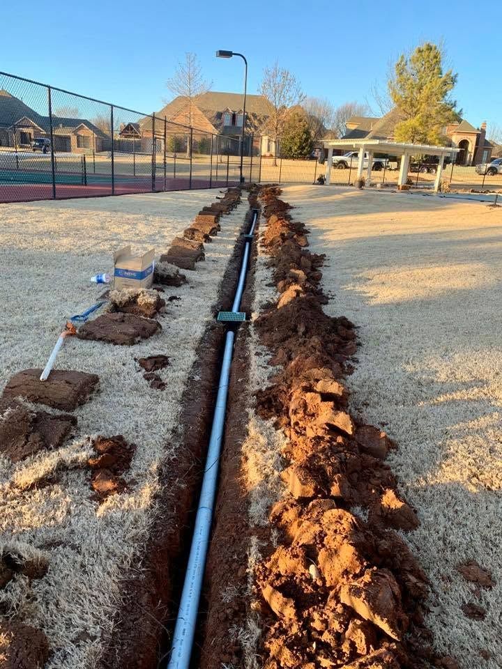 Trenched blue irrigation pipe beside a dirt path in a park, with fencing and houses in the background