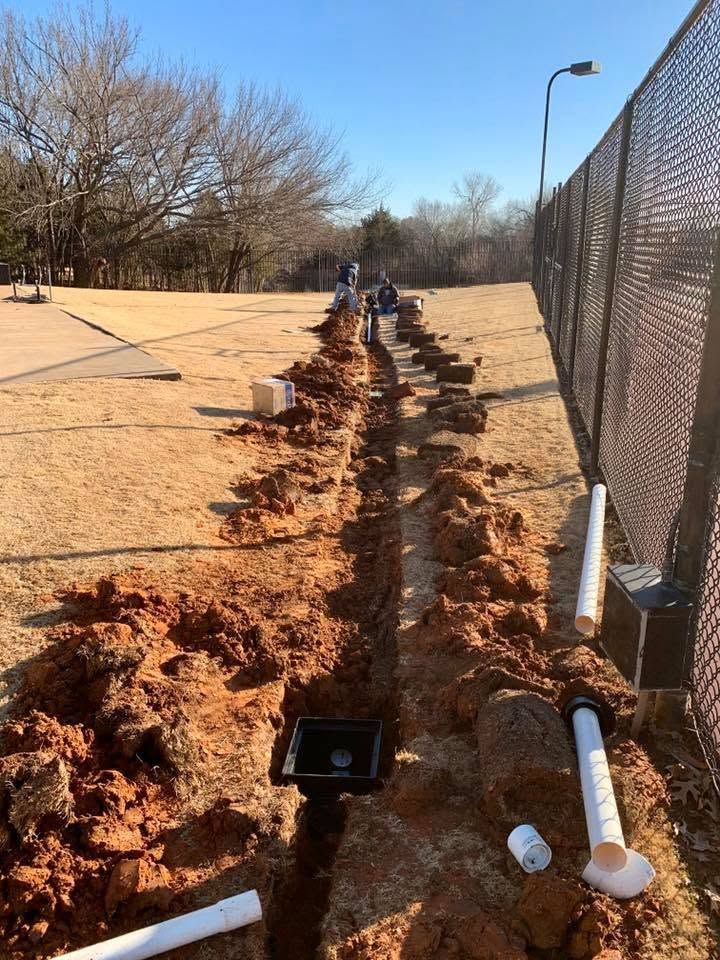 Trenched dirt along a sidewalk beside a chain-link fence, with exposed pipes and workers in the distance.