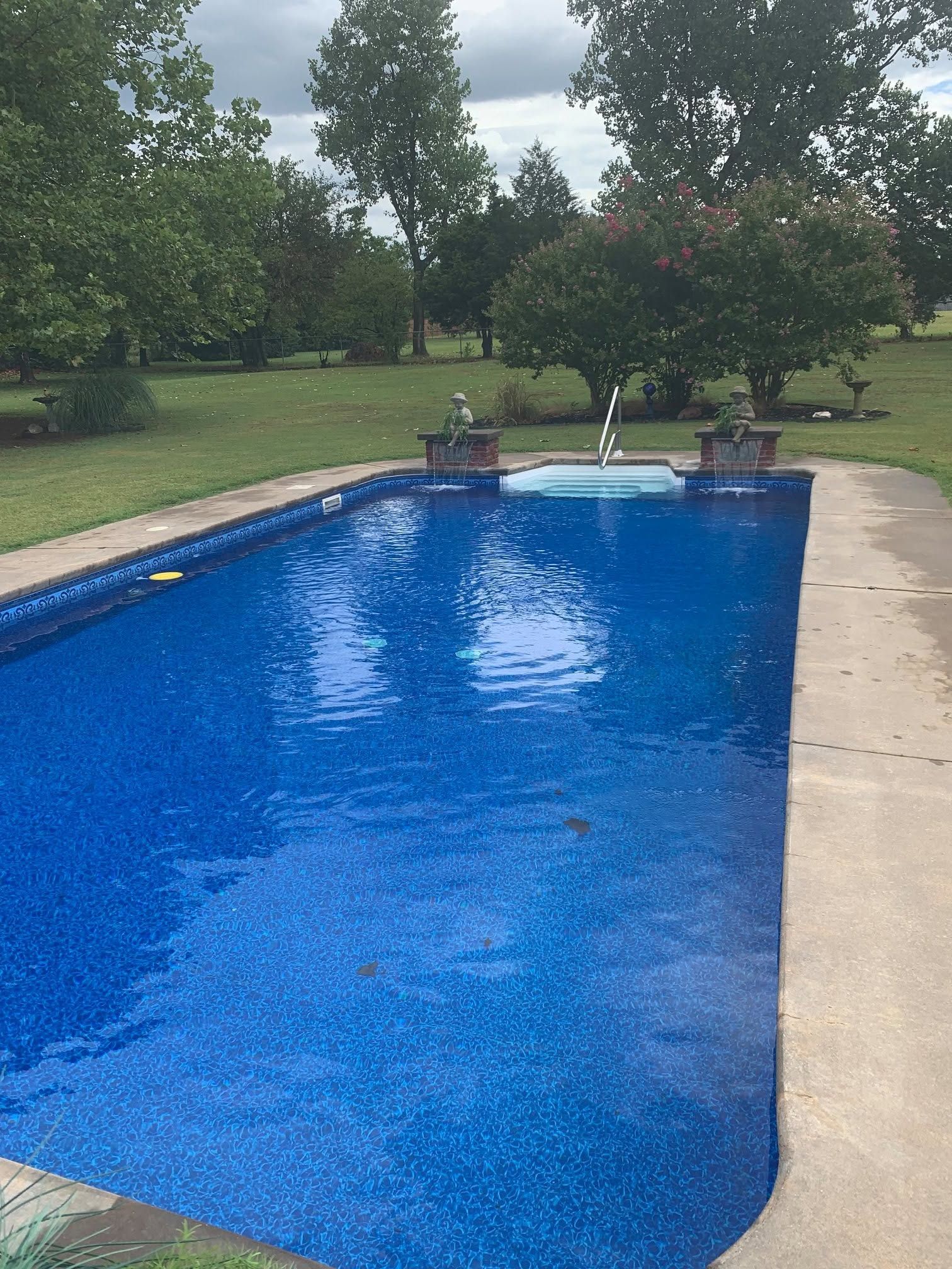 Backyard swimming pool with blue water and surrounding patio, trees in the background