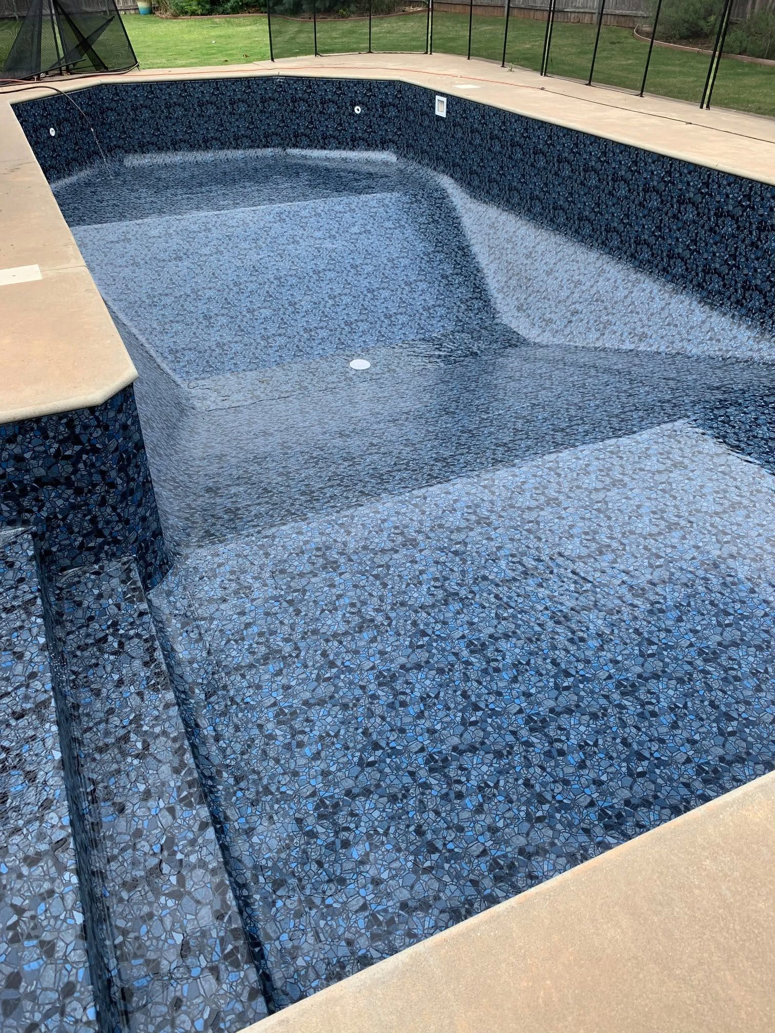Empty in-ground pool with blue-speckled lining, tan deck, and black fence in a backyard.