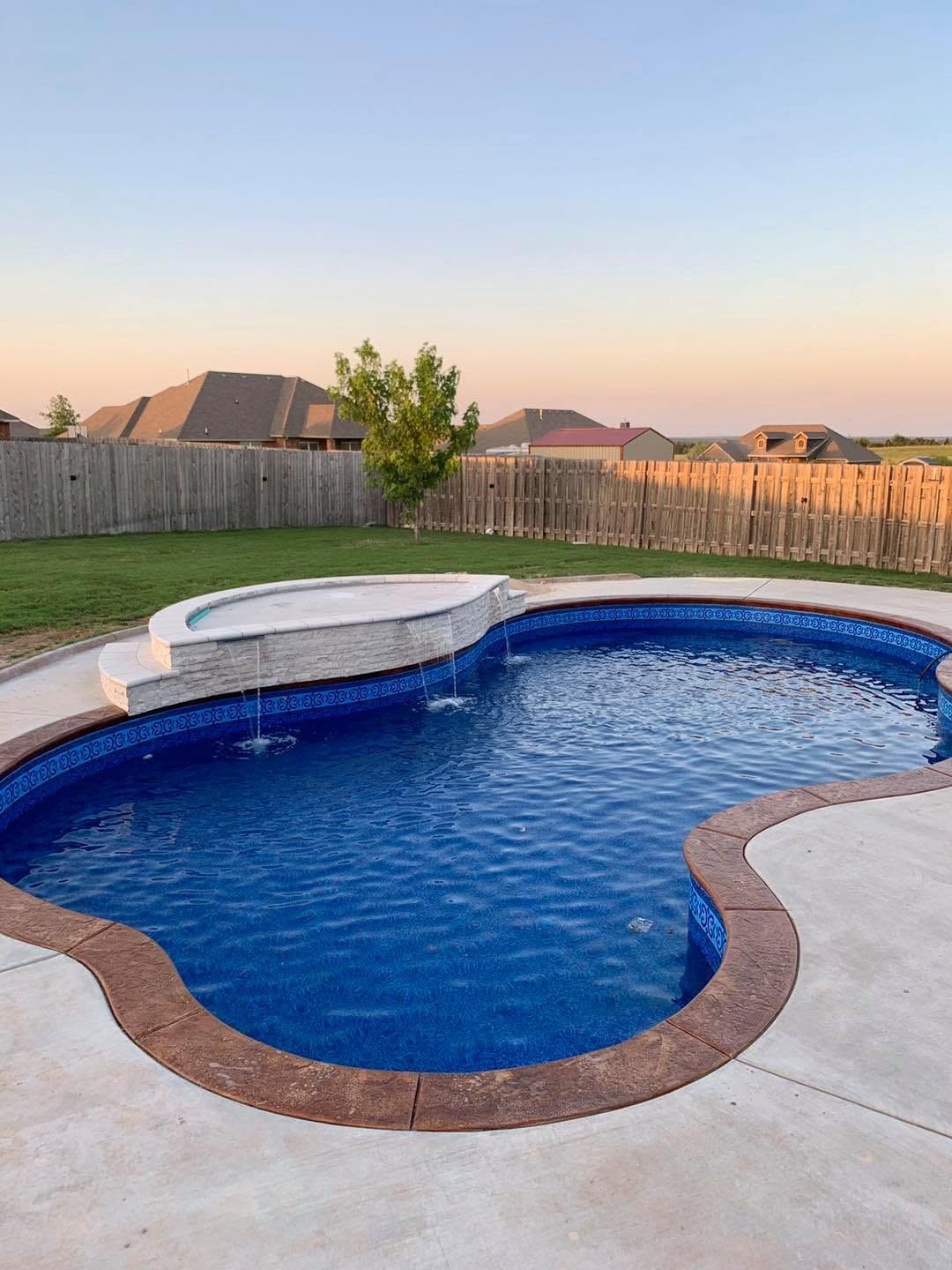 Backyard swimming pool with blue water, white spa ledge, and wooden fence at sunset