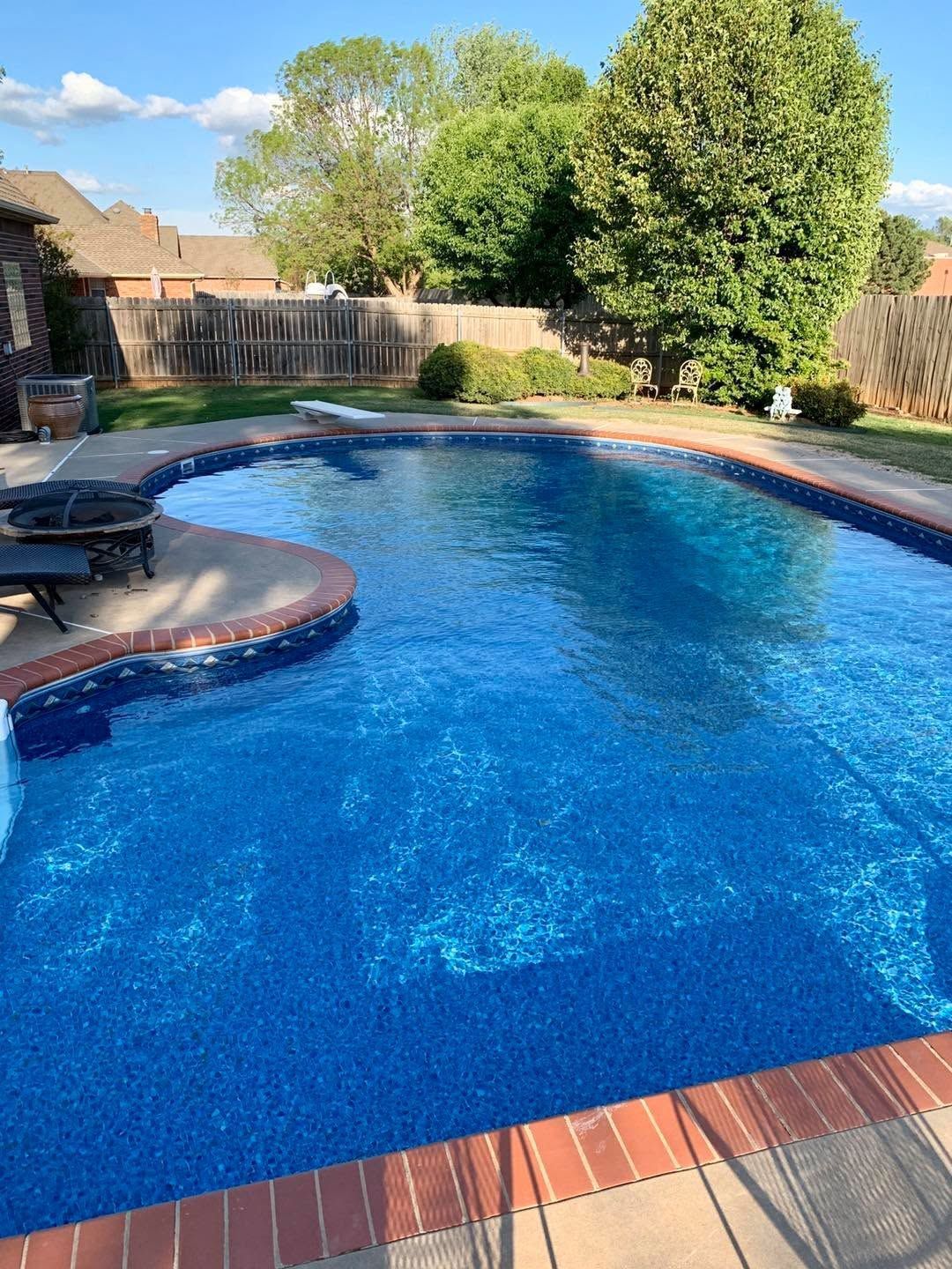 Backyard swimming pool with blue water, brick edge, patio chairs, and a large tree in the background