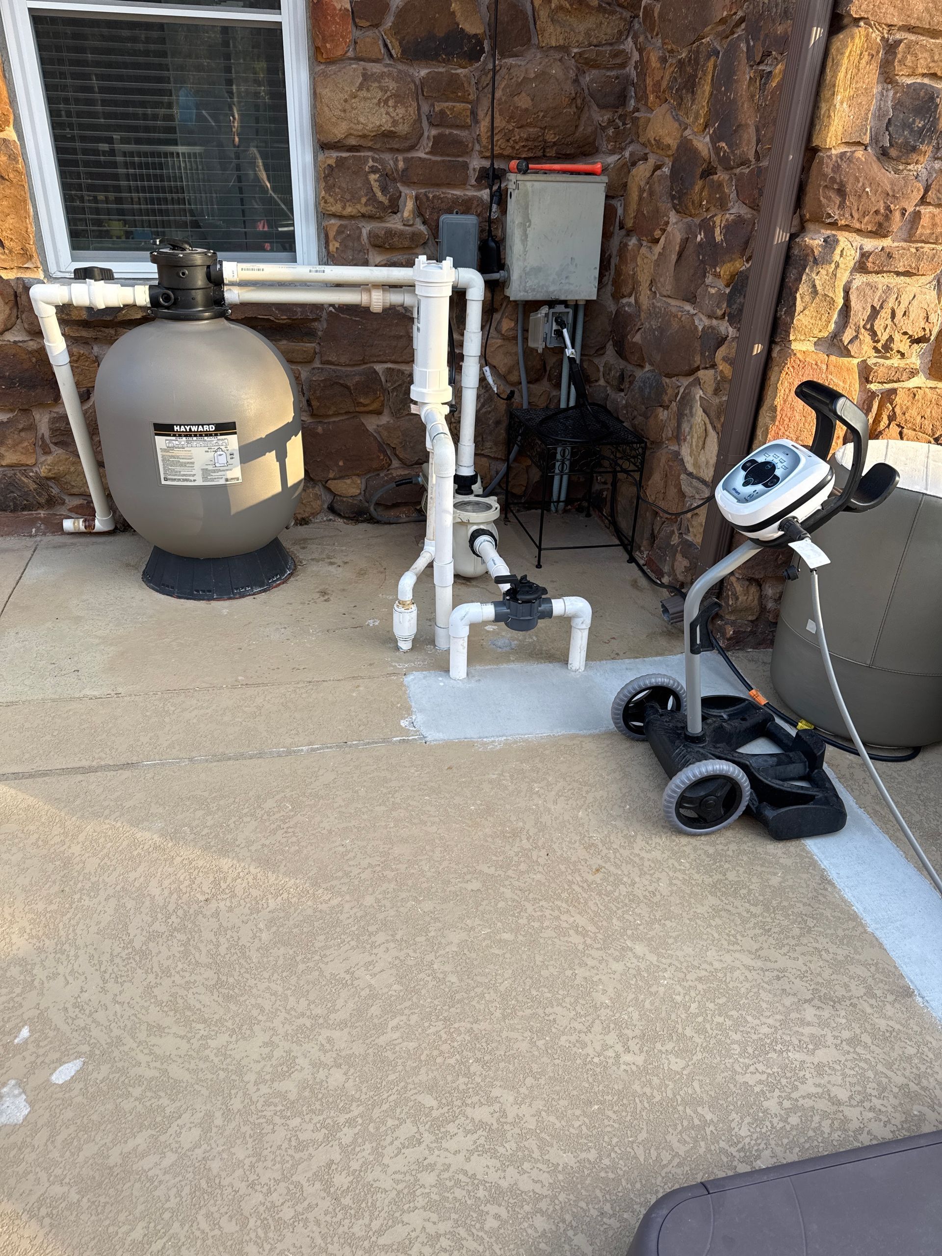 Pool equipment beside a stone wall: a tank, pipes, control box, and a wheeled skimmer on a concrete pad.