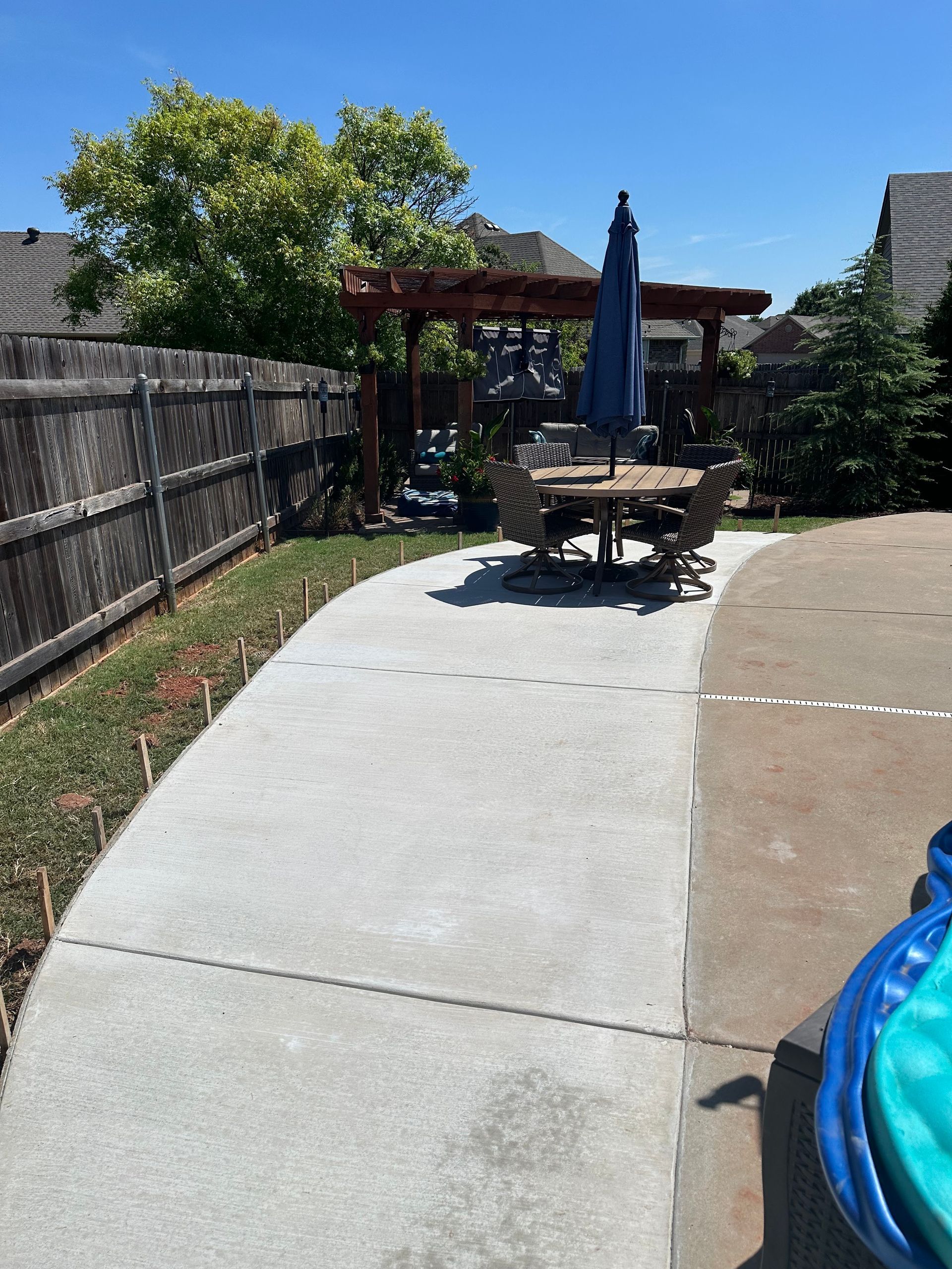 Backyard patio with concrete path, picnic table, blue umbrella, and wooden fence in sunlight