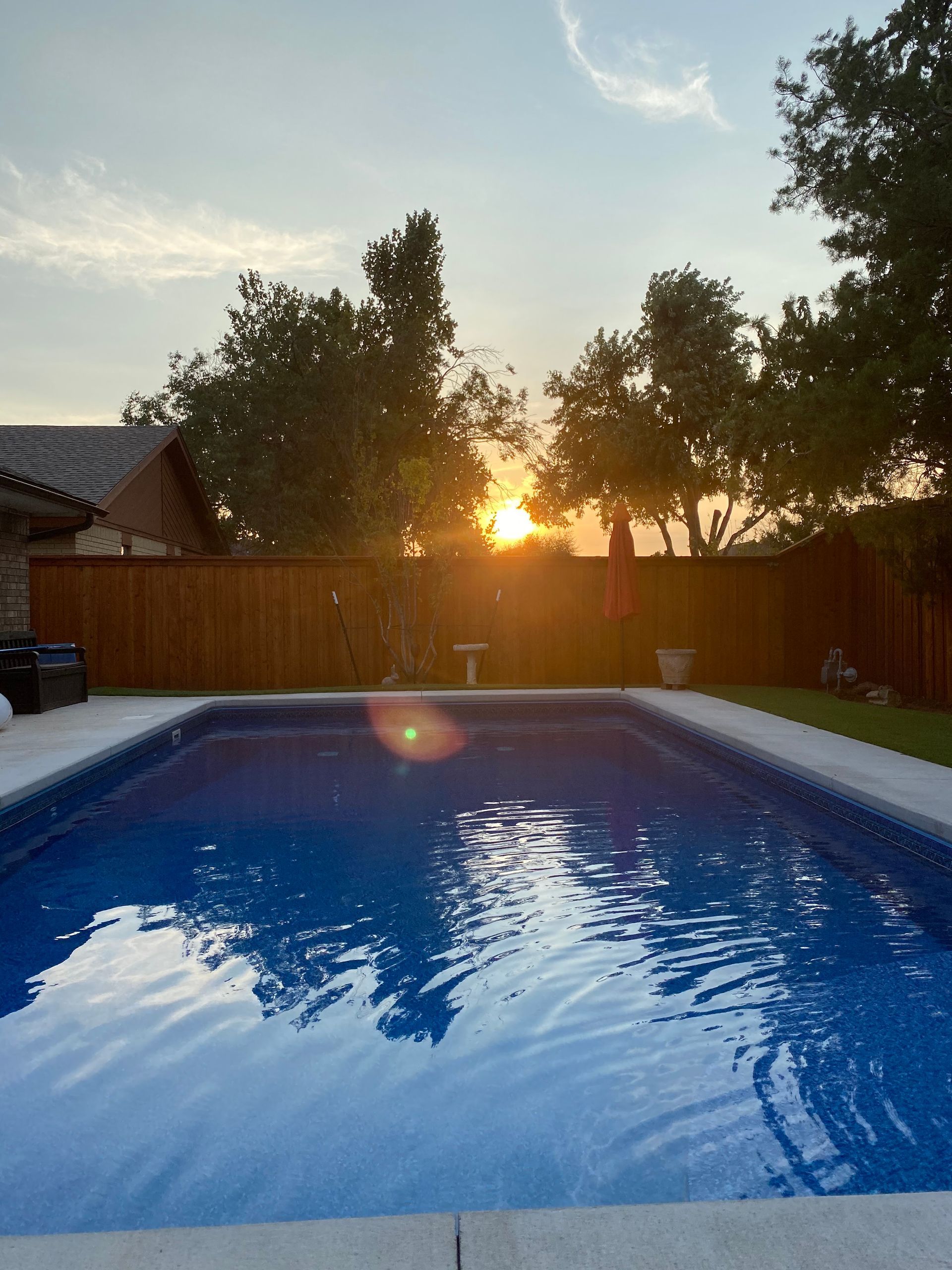 Backyard swimming pool at sunset with trees, wooden fence, and a house in the background