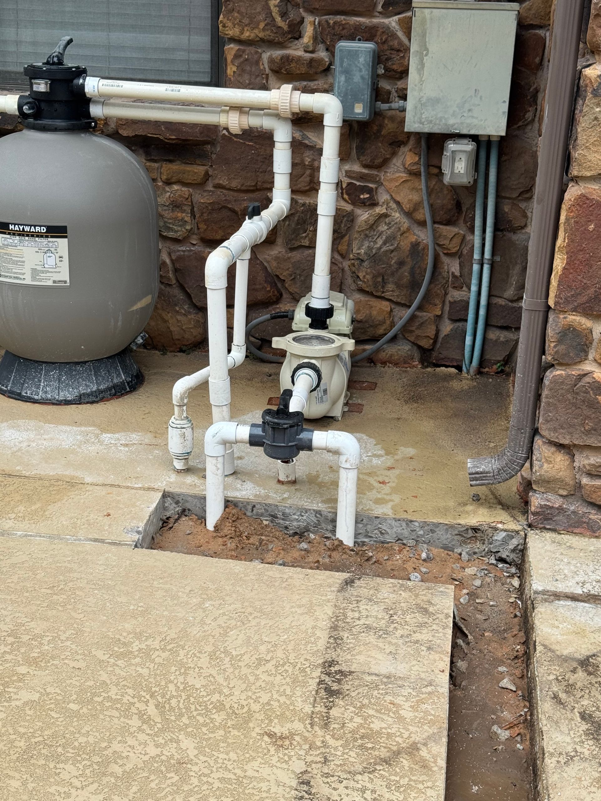 Outdoor pool plumbing with white PVC pipes, valves, and a gray tank beside a stone wall.