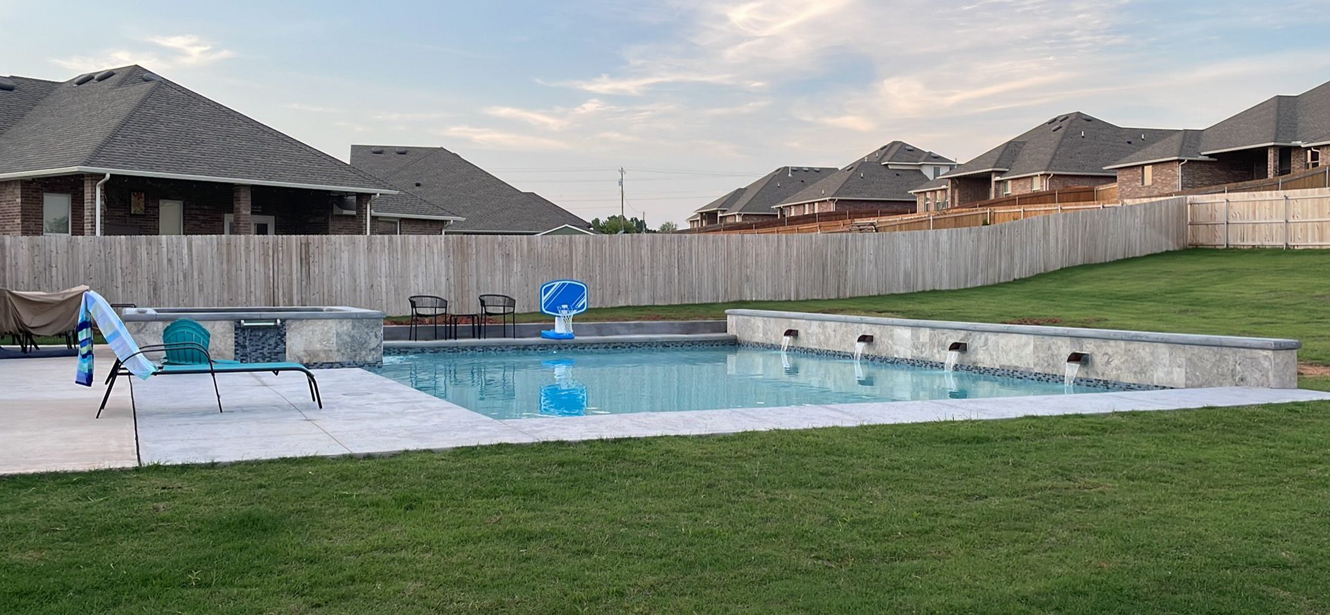 Backyard pool with white deck, blue chairs, lawn, and suburban houses under a cloudy sky
