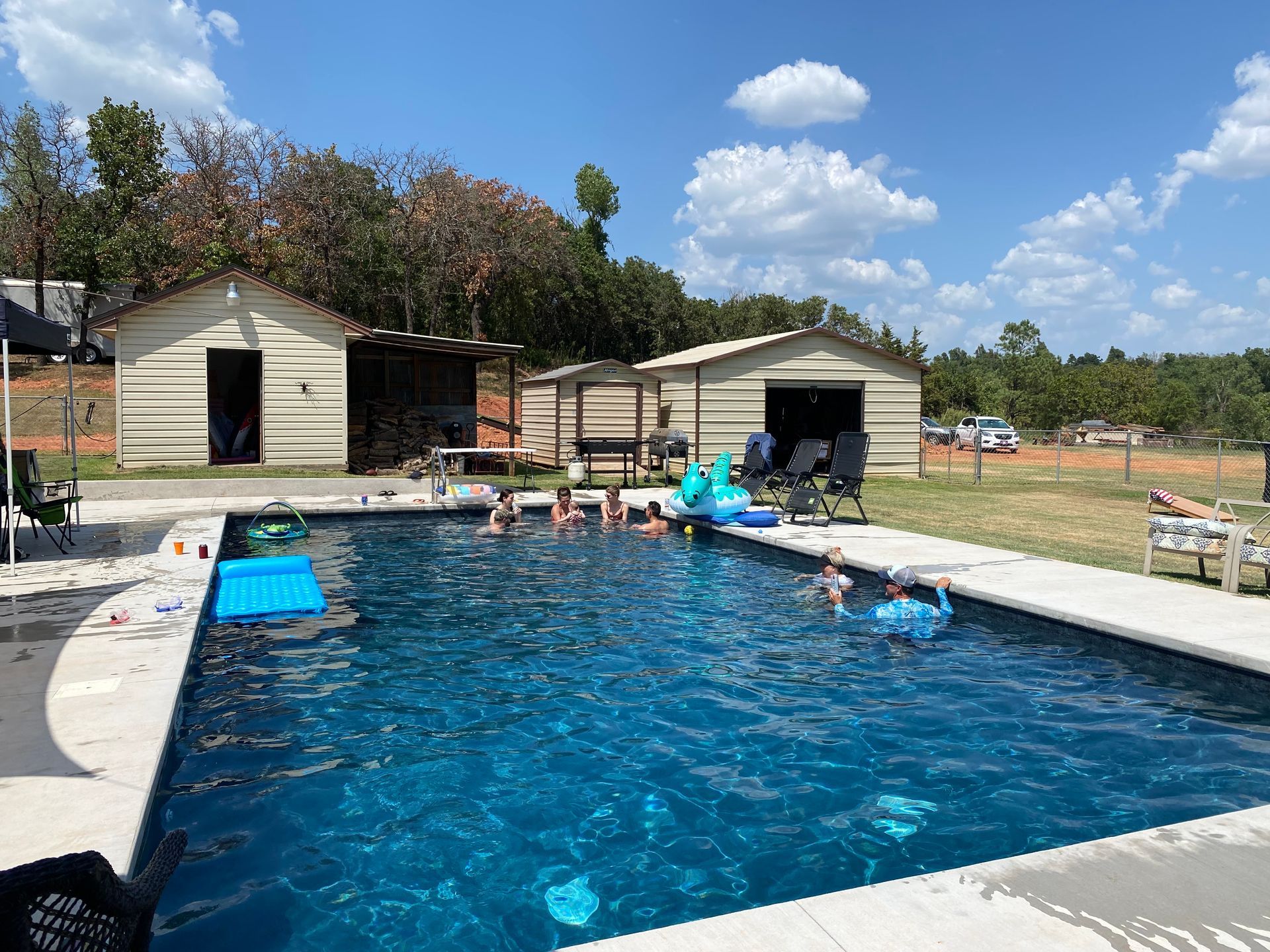 Backyard pool with children’s float toys, lounge chairs, and two sheds under a blue sky