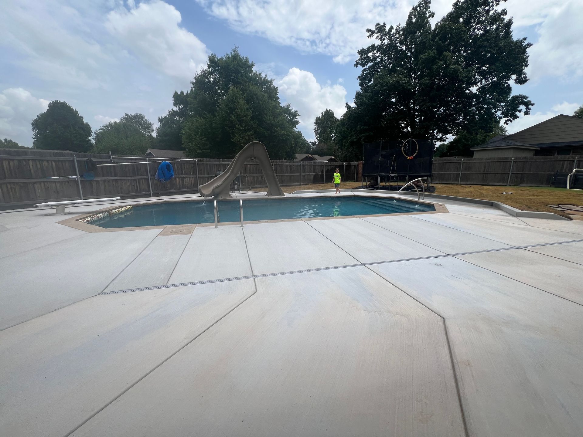 Empty backyard pool under construction with concrete deck, fence, trees, and blue sky