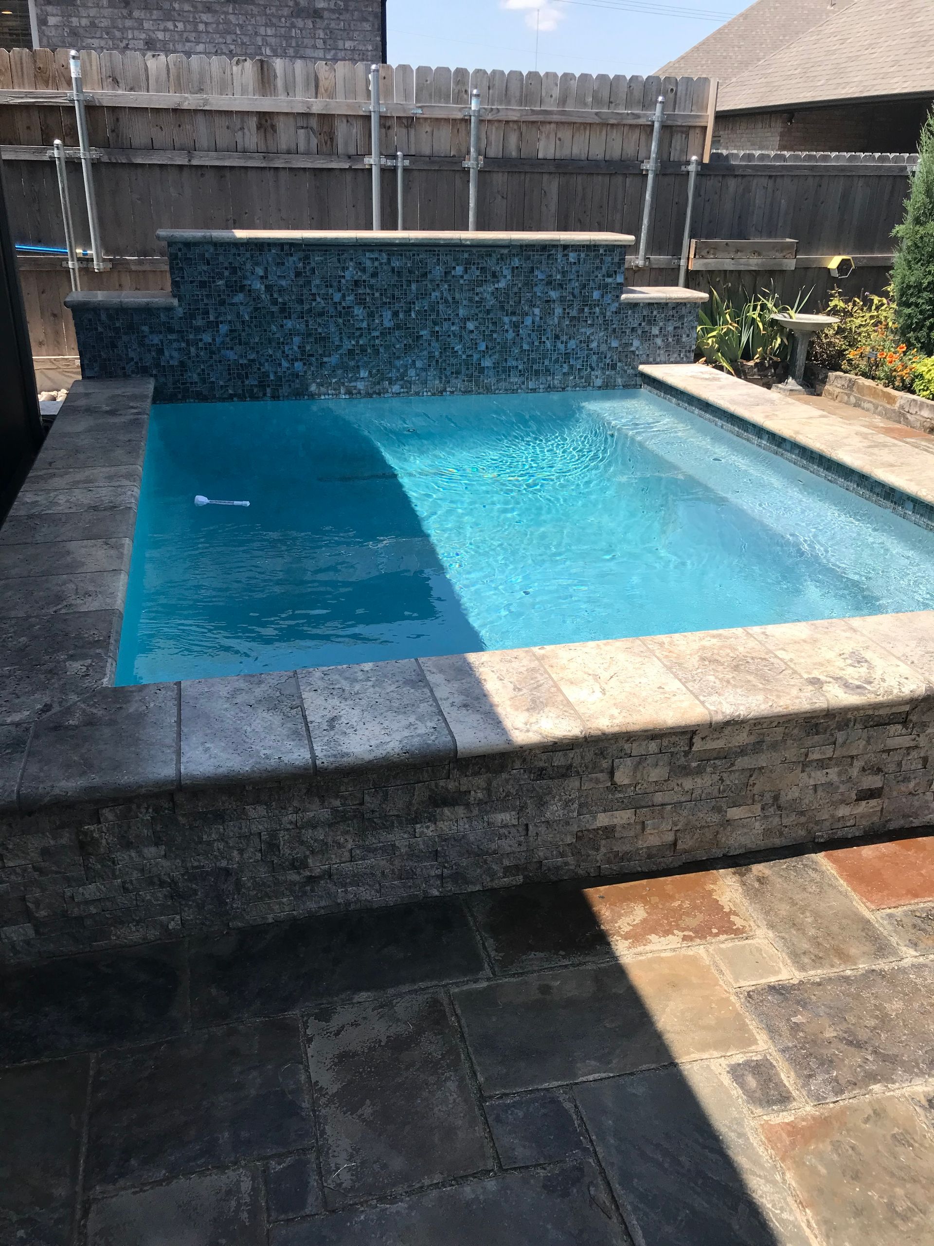 Small concrete backyard pool with blue water and stone patio in sunlight