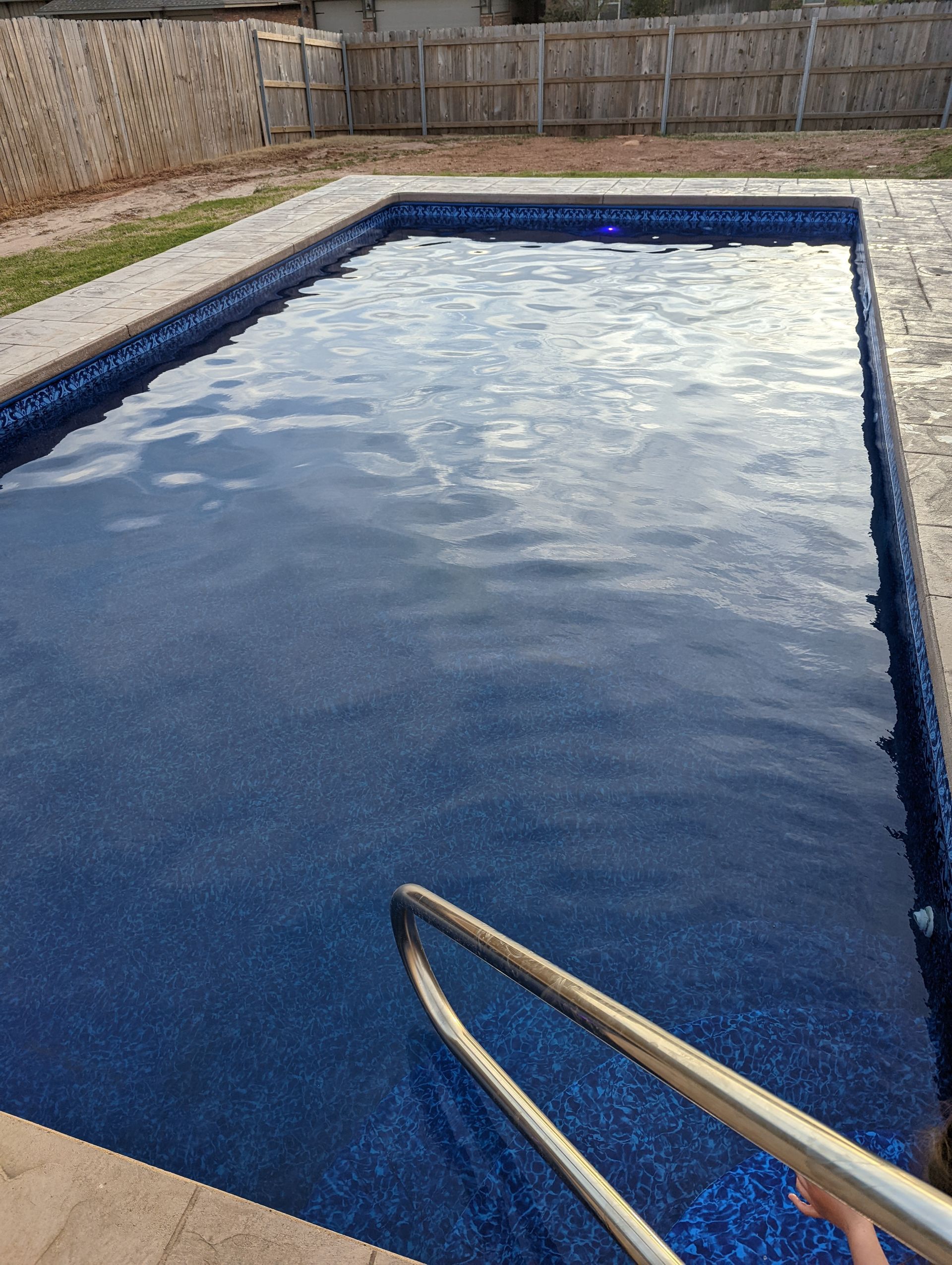 Blue backyard swimming pool with a metal handrail in the foreground and a wooden fence around it