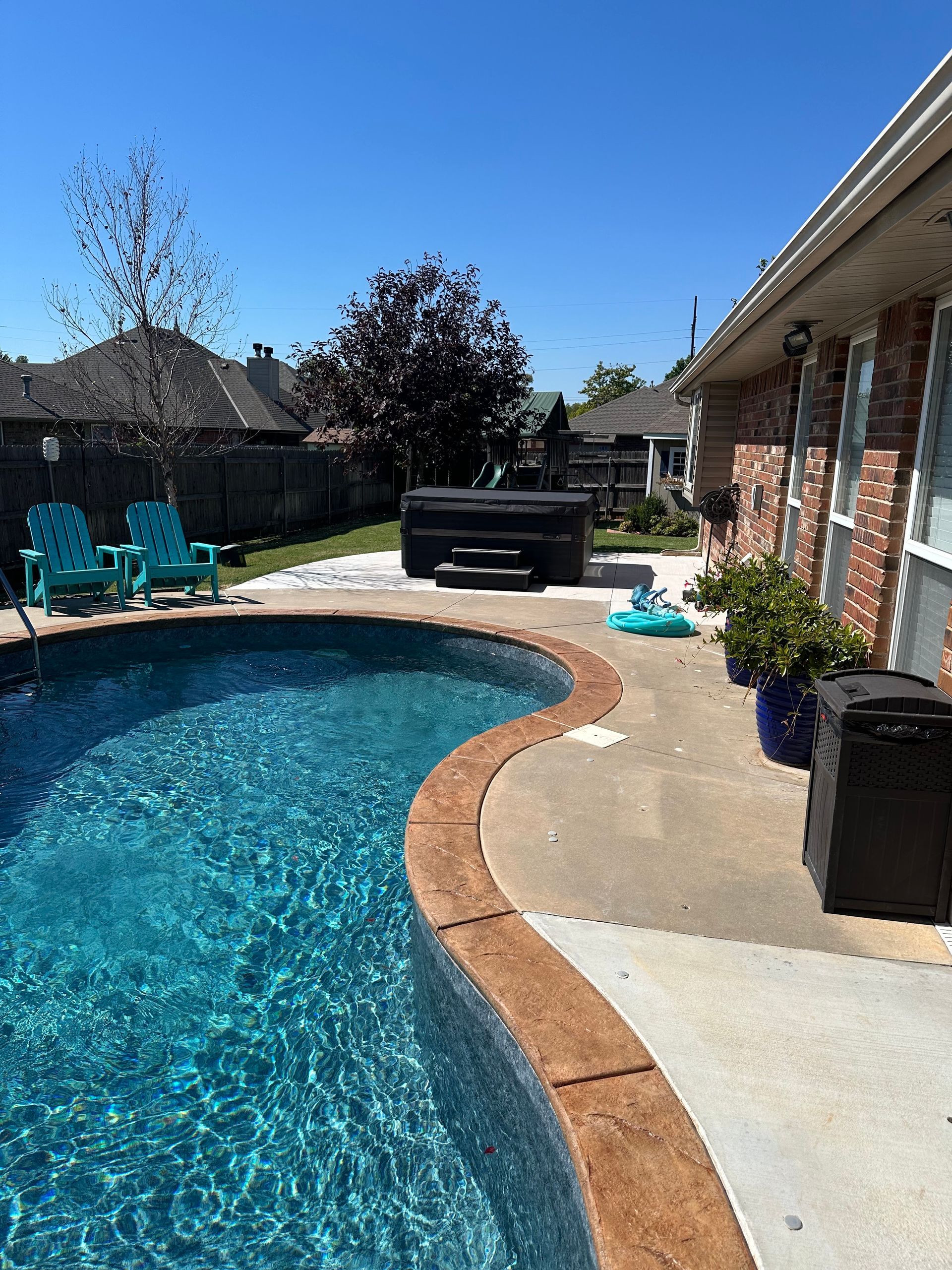 Backyard pool with blue water, patio chairs, hot tub, and brick house in bright sunlight