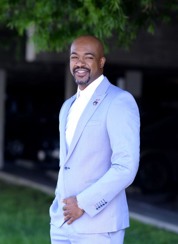 A man in a light blue suit is standing in front of a tree.