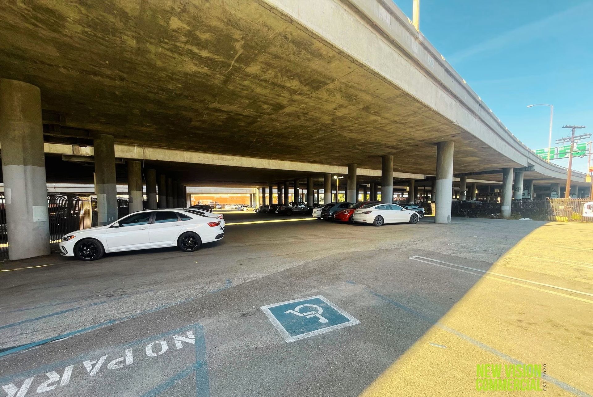 Covered parking area under a highway overpass with several parked cars and blue disabled parking markings