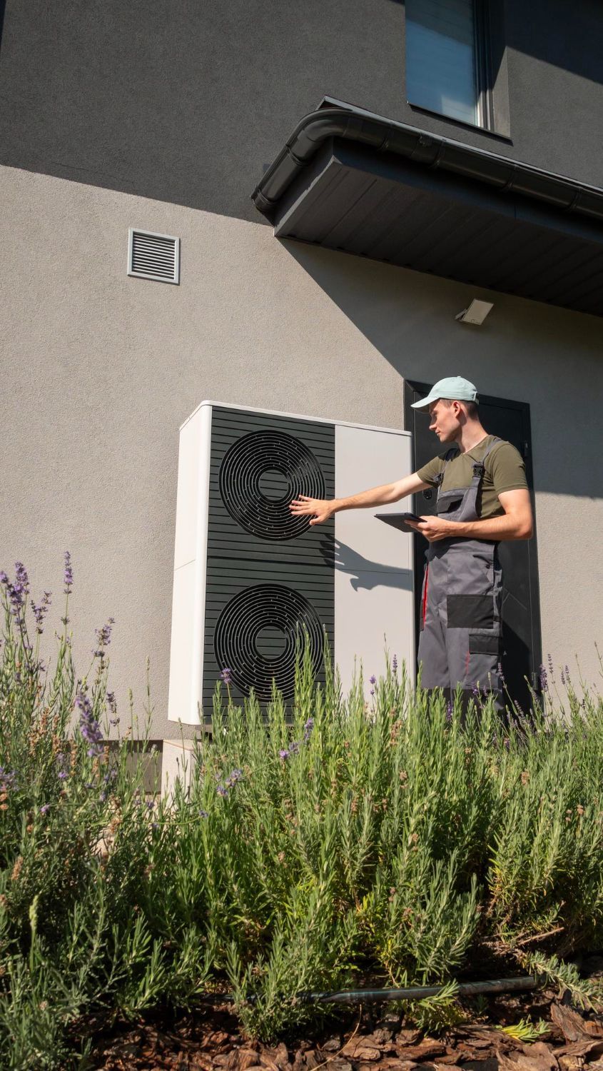 Person in a cap pointing at a white wall-mounted unit outside a modern building, with tall plants in front.