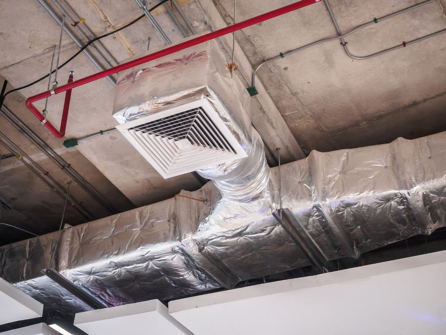 Ceiling air vent with silver ductwork and exposed red pipes in an industrial interior