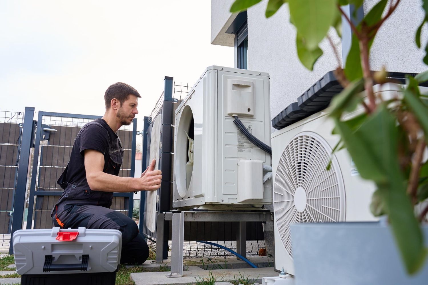 Technician servicing an outdoor air conditioning unit beside a building, with tools and equipment nearby