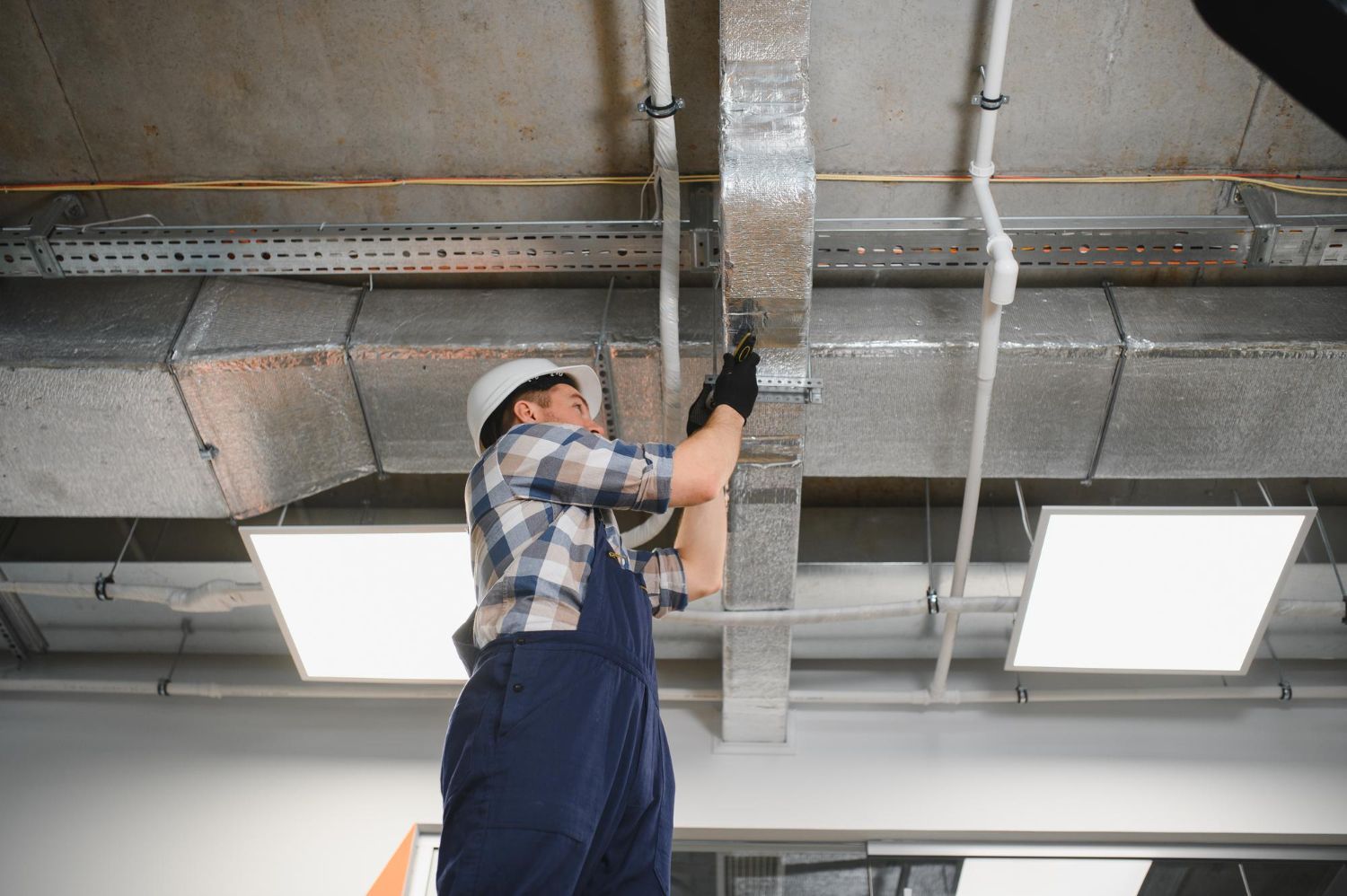 Worker in blue overalls inspects ceiling ductwork with a flashlight in an industrial space