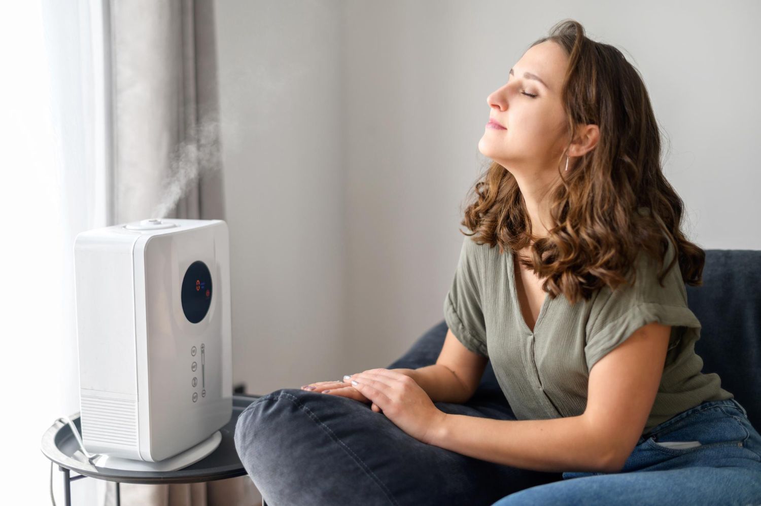 Woman sitting beside a white humidifier, resting on a couch by a bright window.
