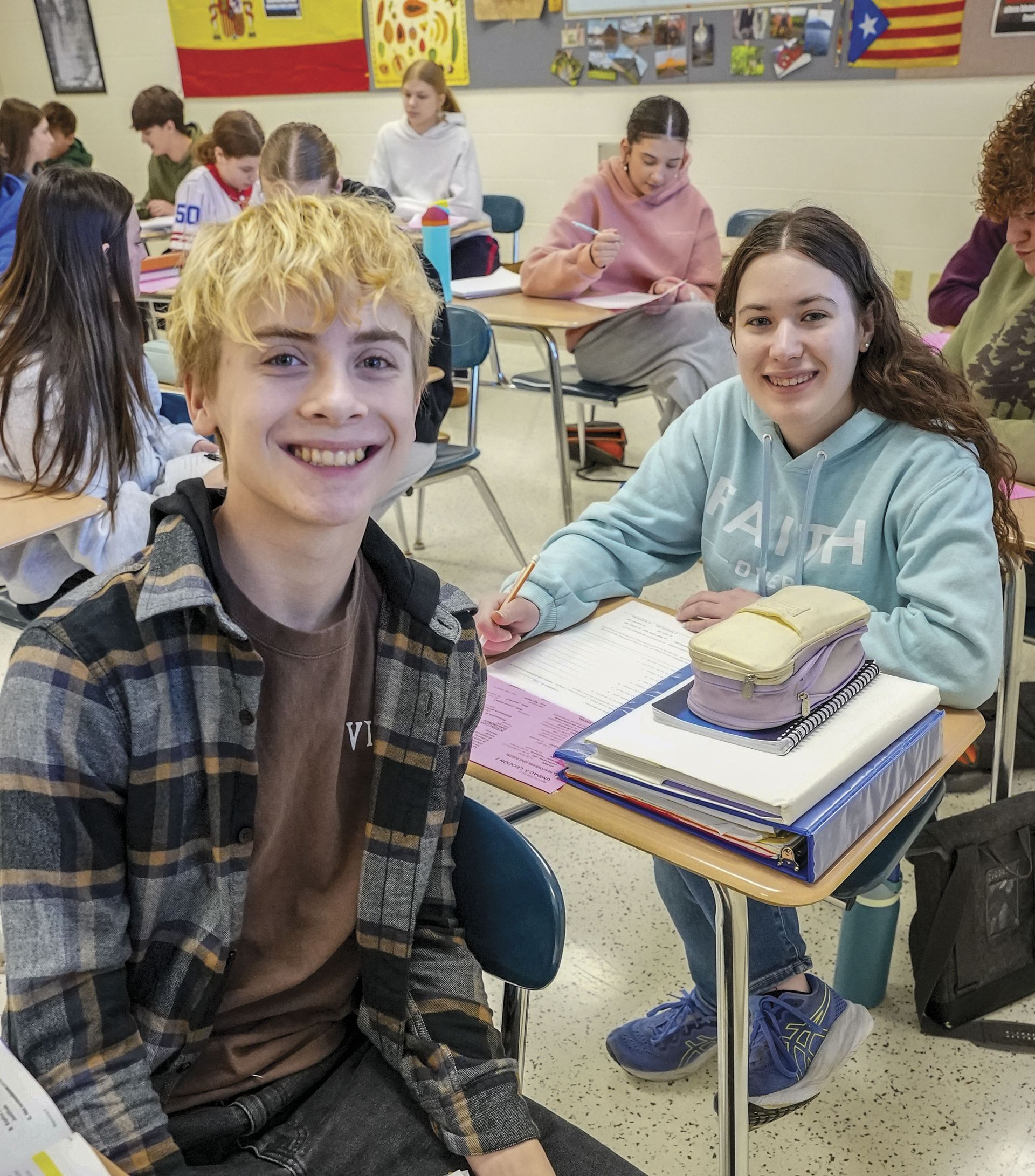 Male and female student in classroom at desks, smiling at camera