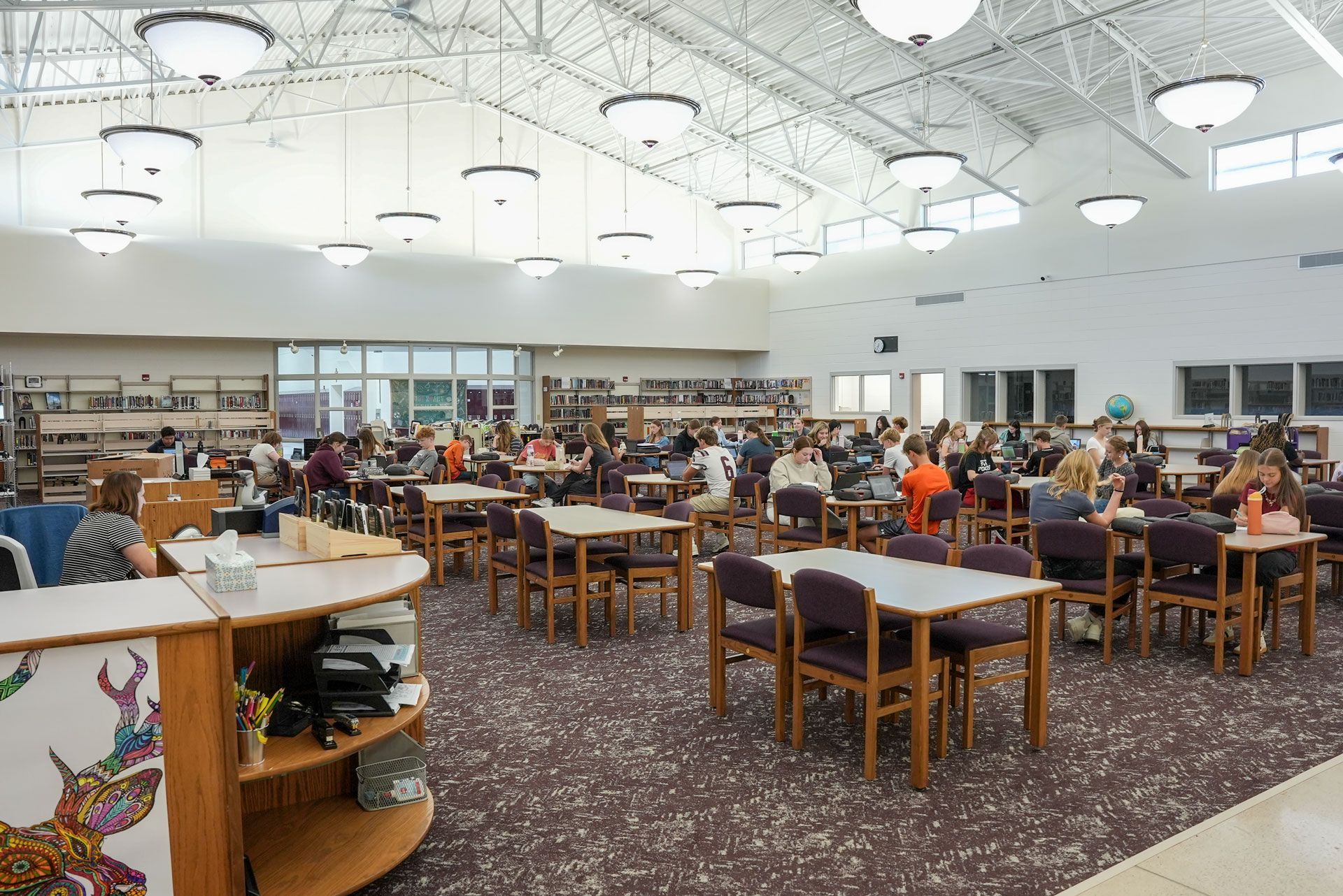 Media Center has high ceilings, many tables and chairs, dark carpeting, and students working at the tables.