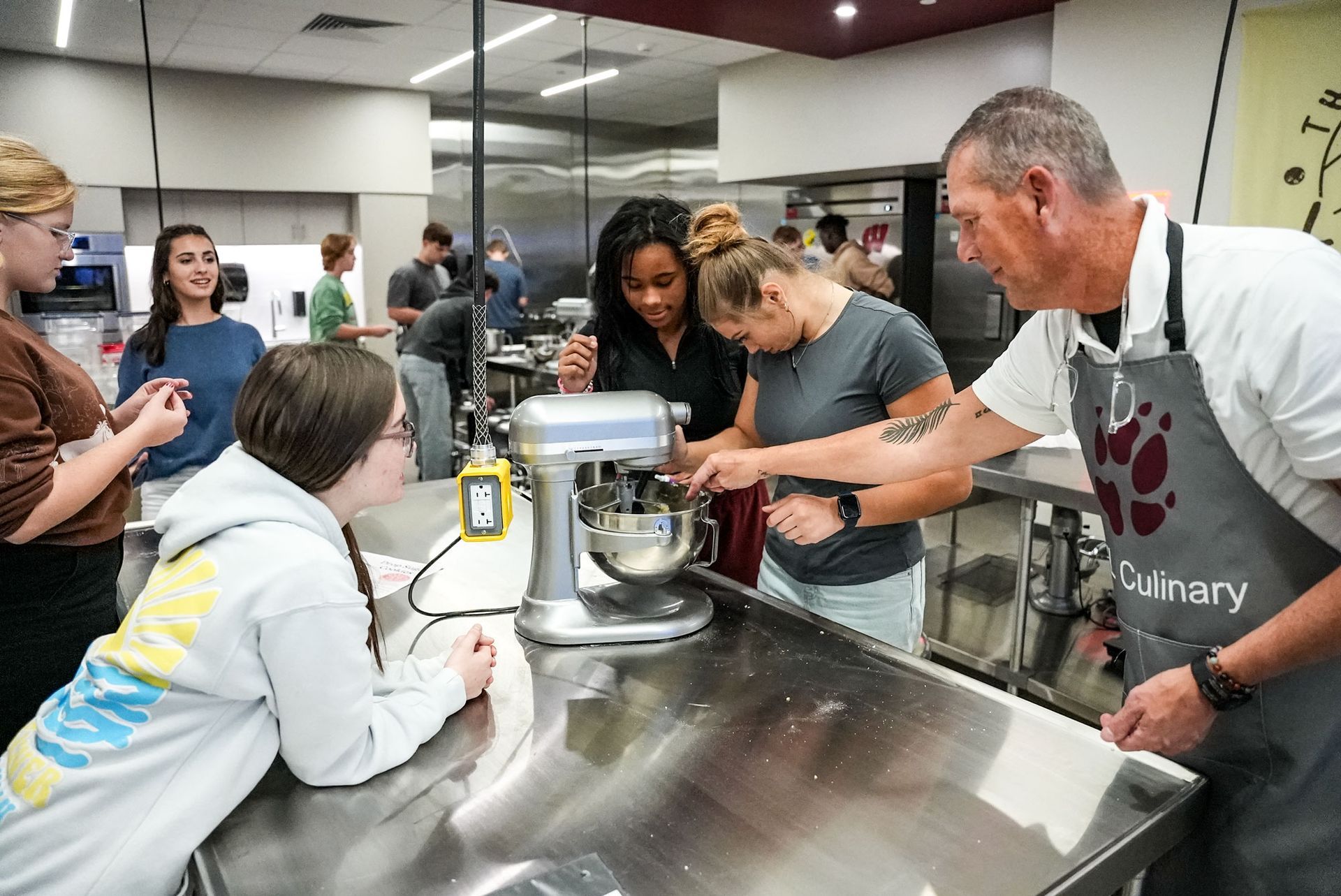 The culinary arts instructor is helping students while they work with a mixer at their station.