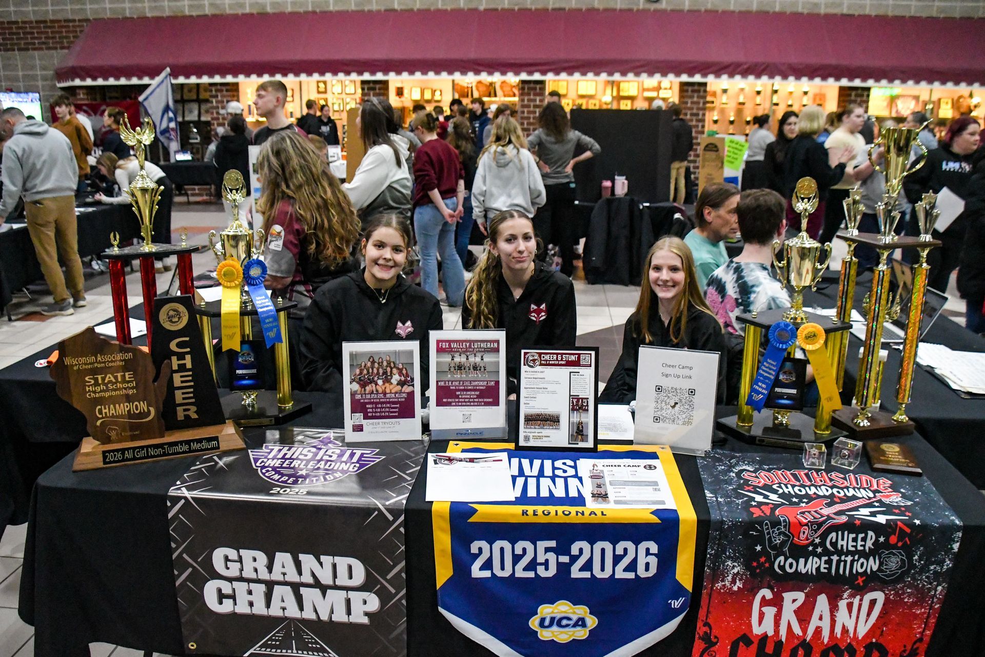 Cheer and stunt table with three smiling girls, trophies, and banners
