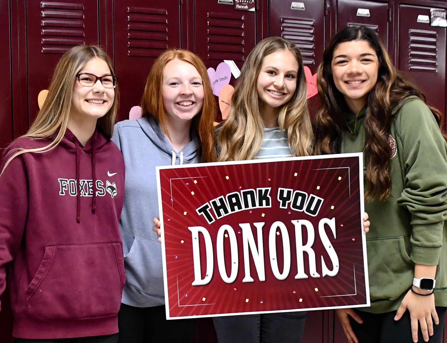 Four smiling girls standing in front of lockers, holding a sign 