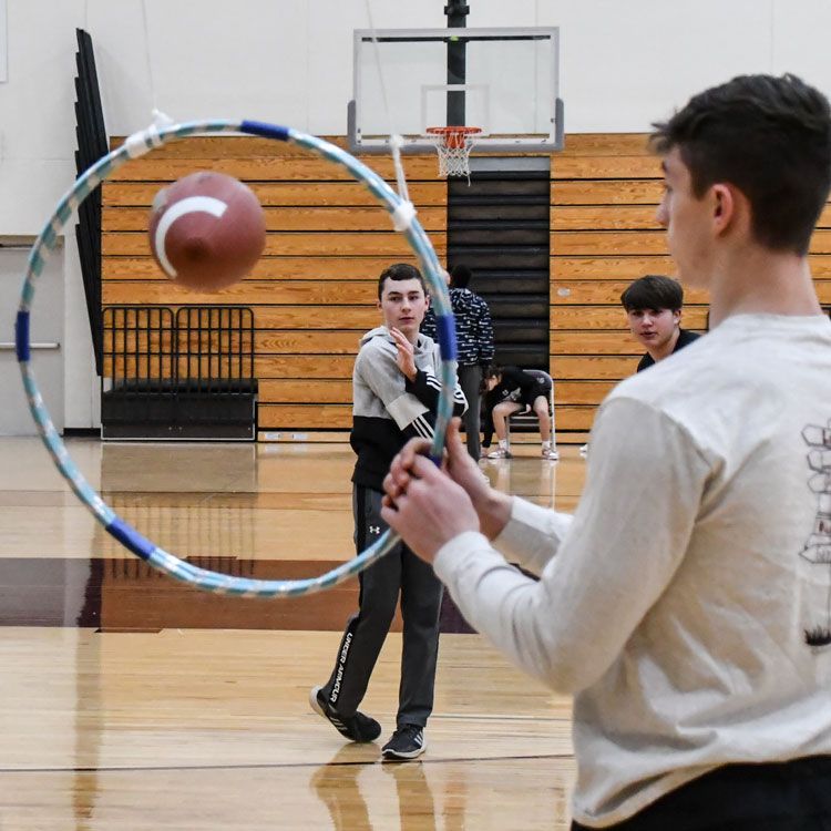 In the gym, one boy holding hula hoop steady while another boy tosses football through the opening