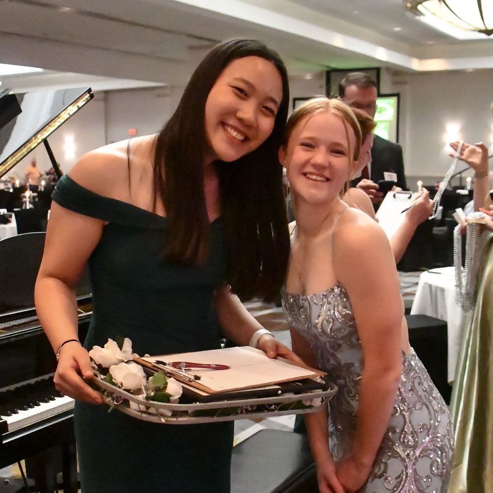 Two smiling high school girls in formal dresses, and holding  a tray of roses for the Rose Lapel event