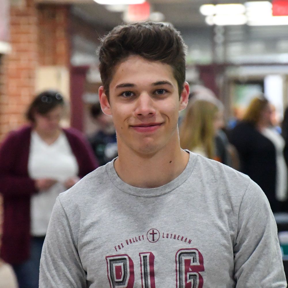 Friendly male student wearing a Peer Leadership Group shirt. He is just inside the busy Commons entrance of school
