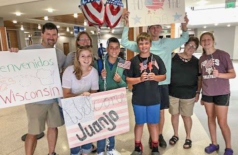Happy group of 8 people at the airport, with balloons, signs, and small USA flags.